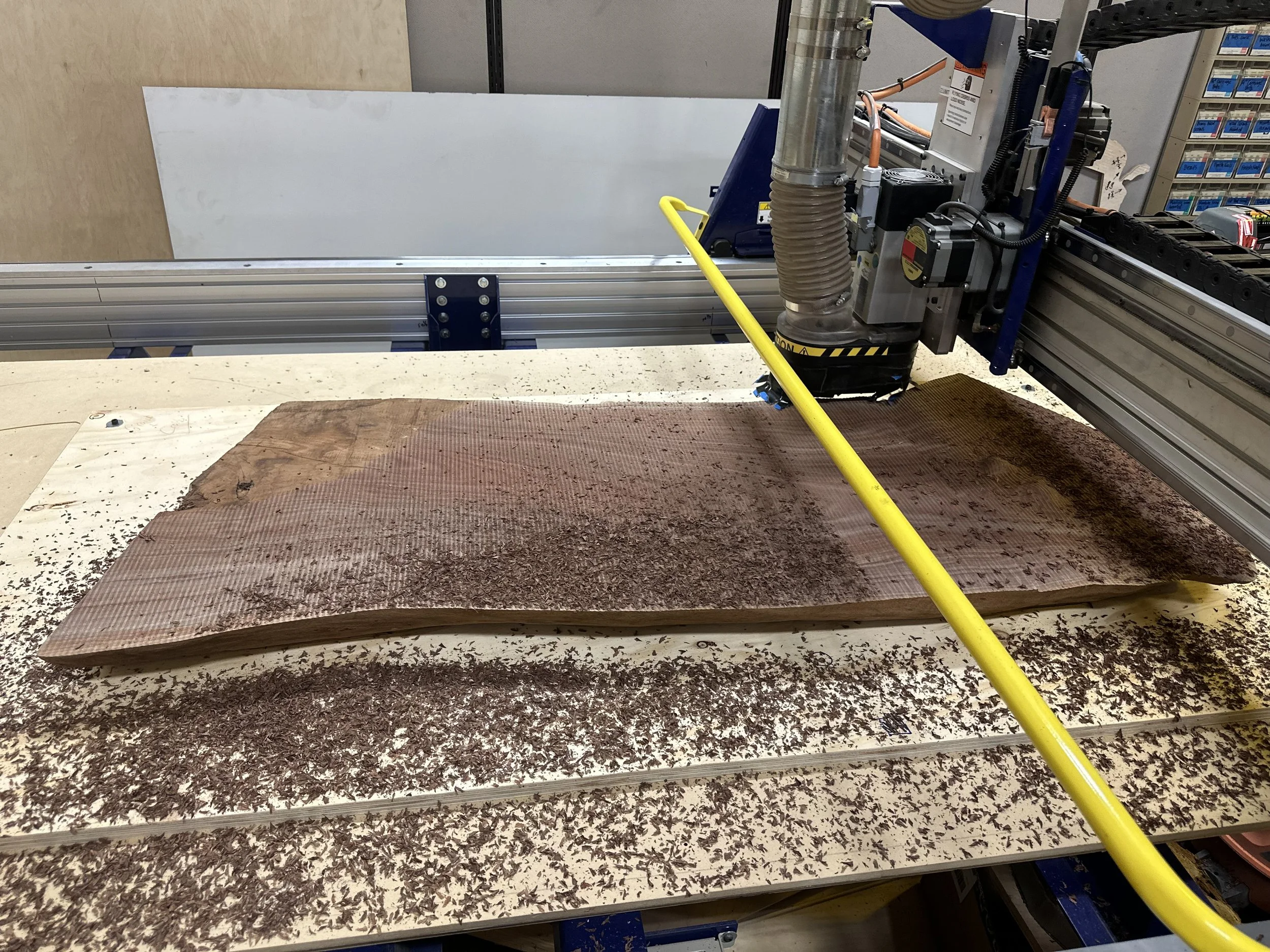 A CNC router machine cutting a piece of wood, with wood shavings scattered on the table beneath.