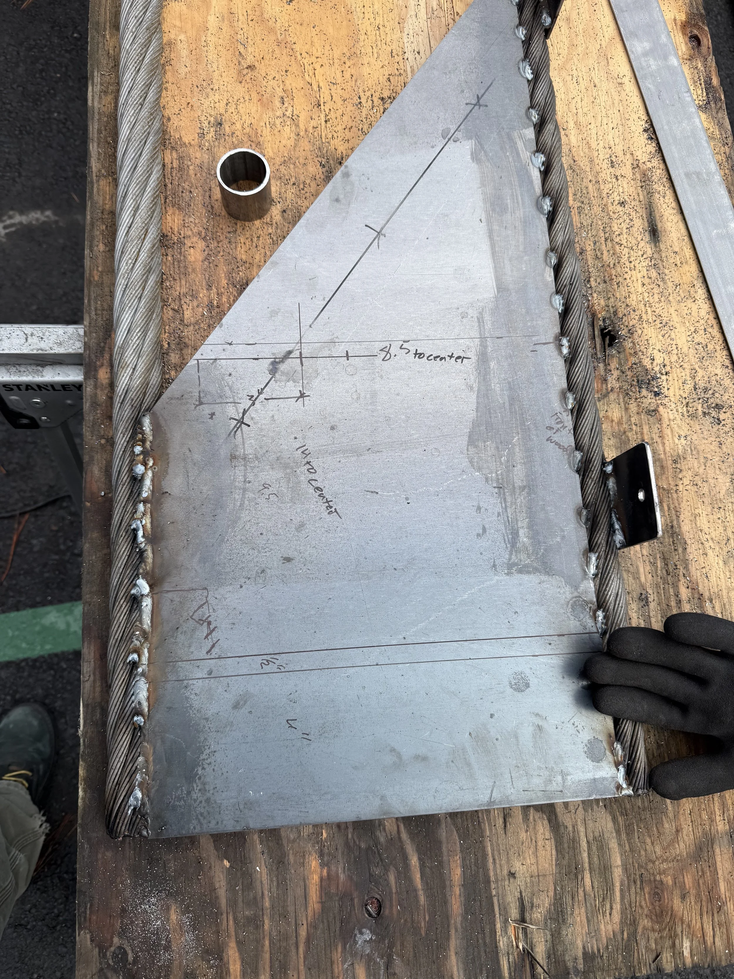 A metal welding project on a wooden workbench, featuring a drilled metal plate with handwritten measurements, a twisted steel rebar frame, and a gloved hand.