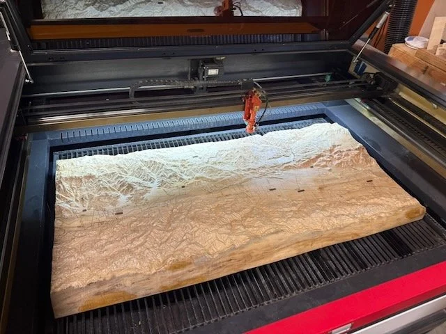 A laser cutter engraving a detailed topographical map of a mountainous landscape on a large piece of wood.