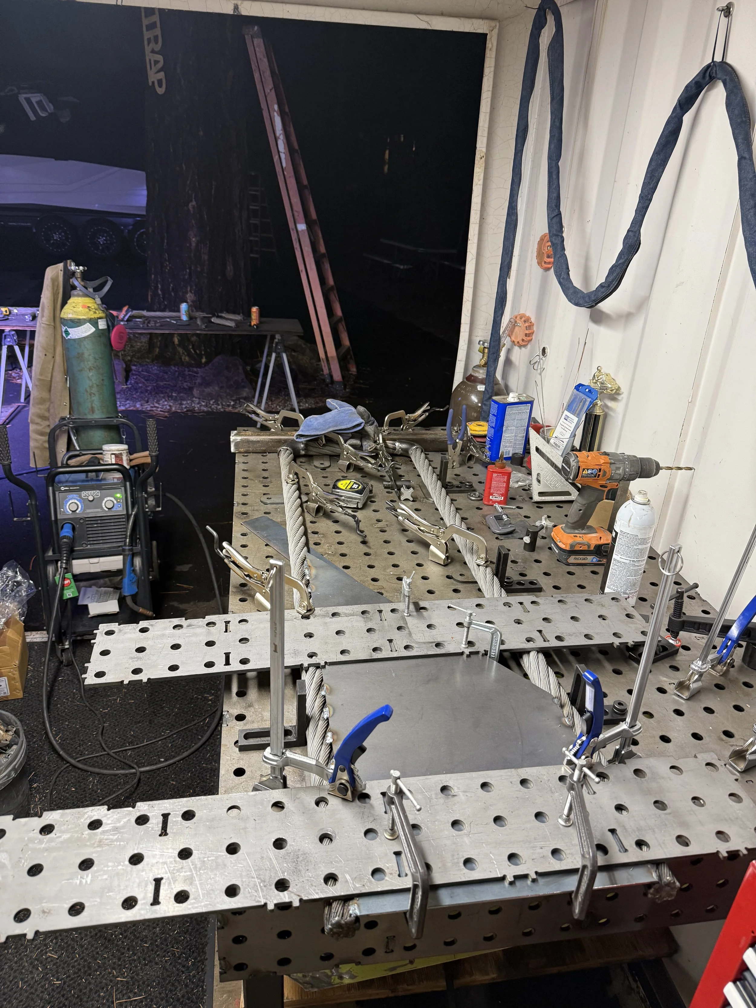 Workshop workspace with tools, clamps, a cordless drill, safety gear, and a large metal work table with a pegboard surface, with a backdrop of a forest scene on a wall.