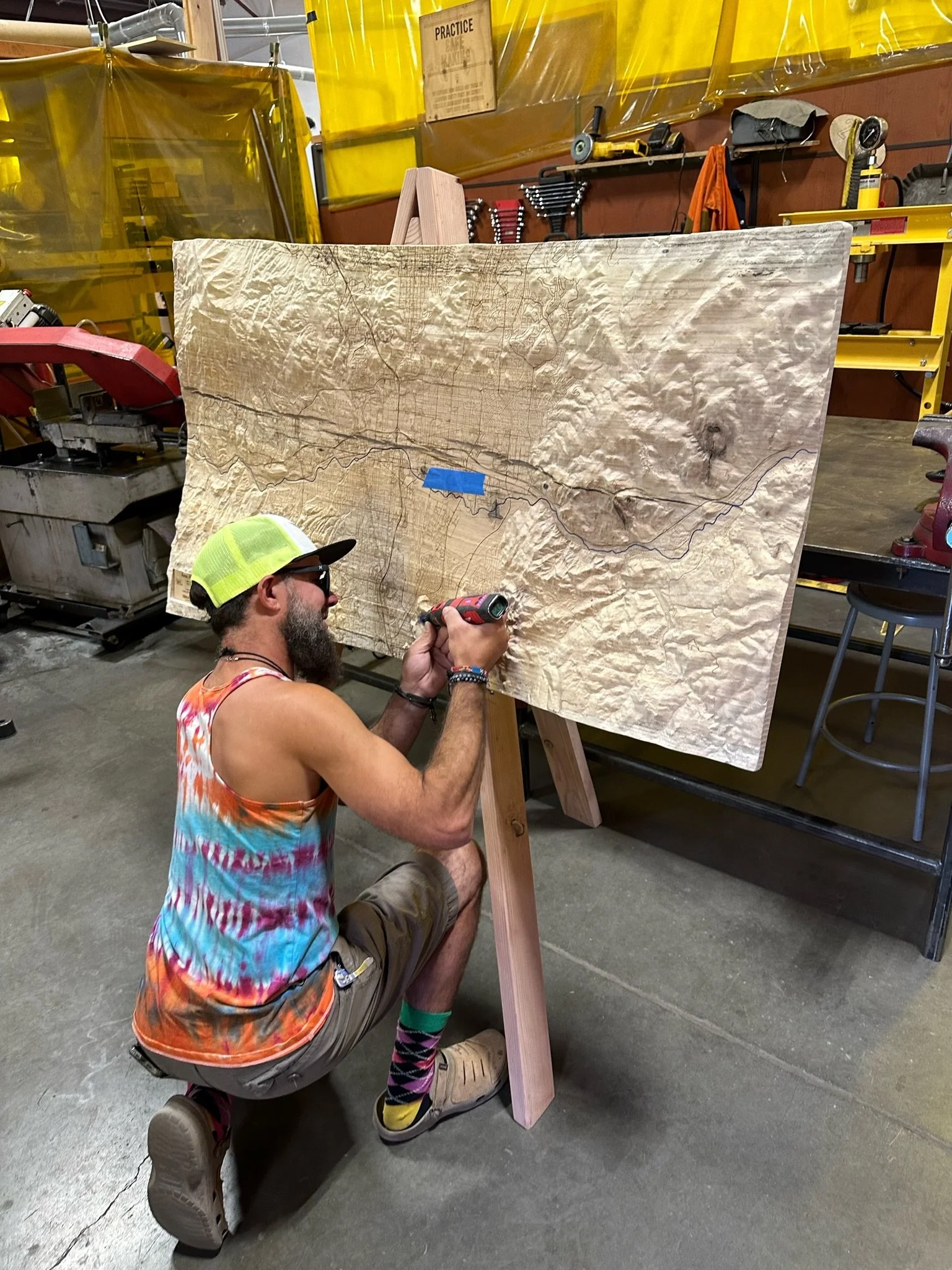 Man with a yellow cap, rainbow tie-dye tank top, and tan shorts kneeling and using a drill on a large, textured wooden map in a workshop with tools and yellow safety barriers in the background.