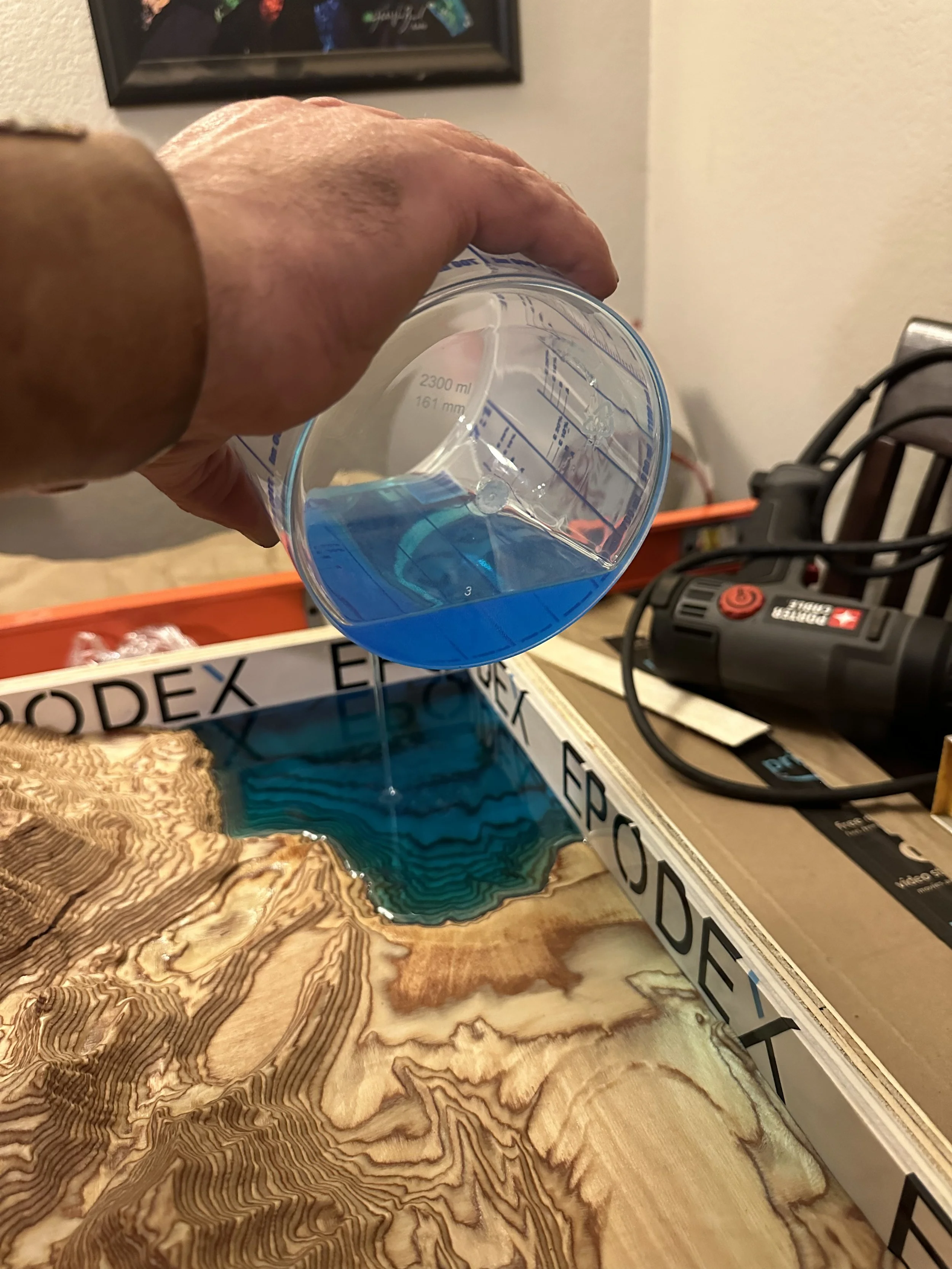A person pouring blue epoxy resin onto a wooden surface with a grain pattern for a tabletop project.