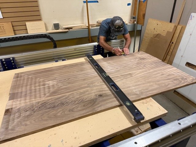 Person working on a large wooden board in a woodworking shop, with a level placed on the board and woodworking supplies and tools in the background.