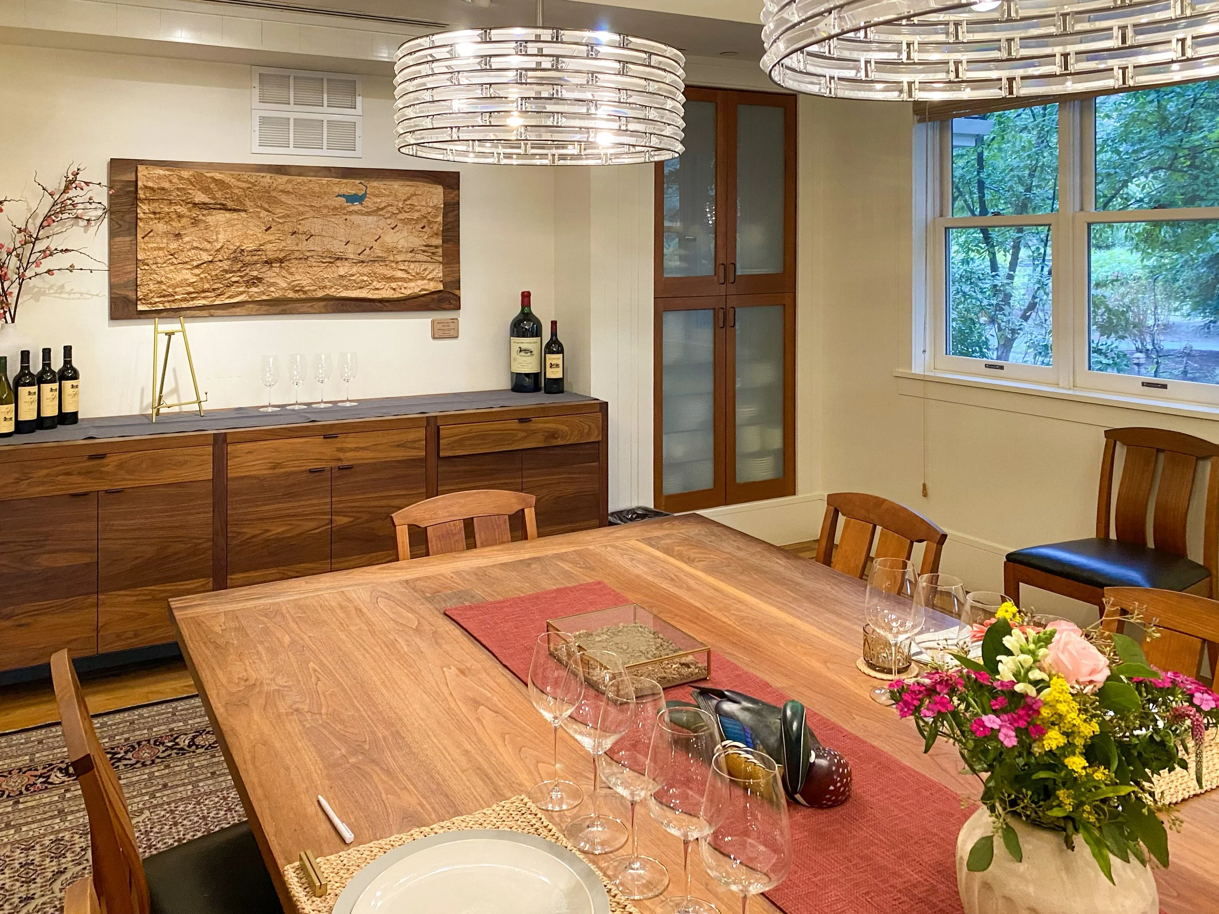 A dining room with a wooden table set with wine glasses, a floral centerpiece, and a place setting. There is a sideboard with wine bottles and glasses, decorated with a branch with red berries. The room has large windows showing green trees outside, and artwork on the wall.