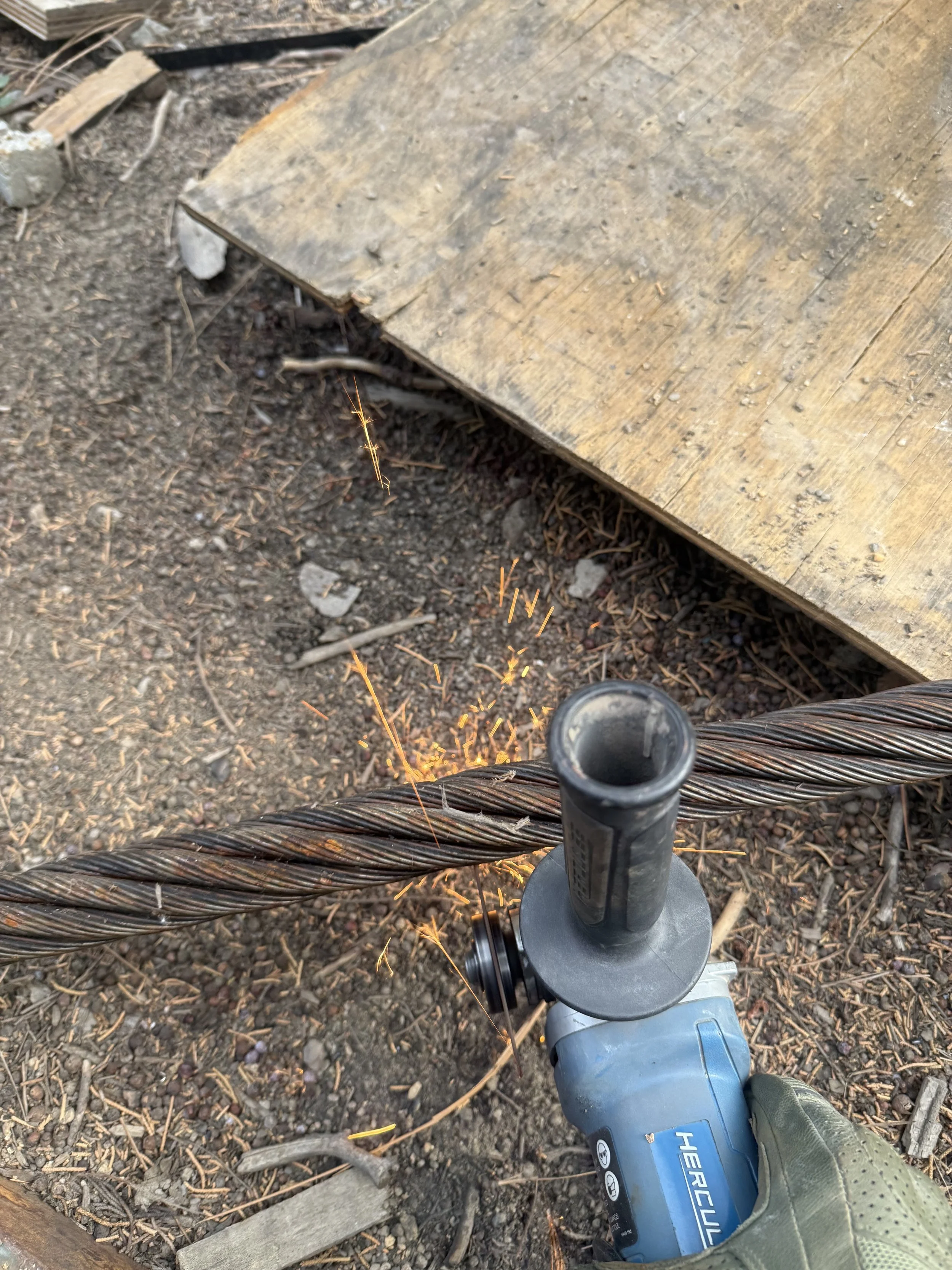 A person using an angle grinder to cut or grind a steel wire rope, with sparks flying, outdoors on dirt ground near a wooden platform.