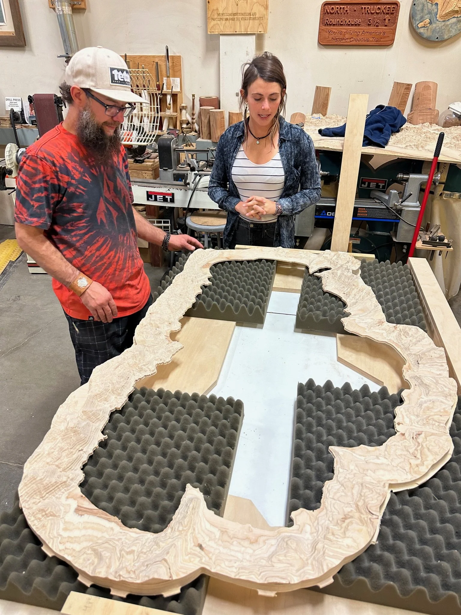 Two people in a woodworking workshop observing a large, irregularly shaped wooden piece with foam padding underneath, possibly for soundproofing or display purposes.