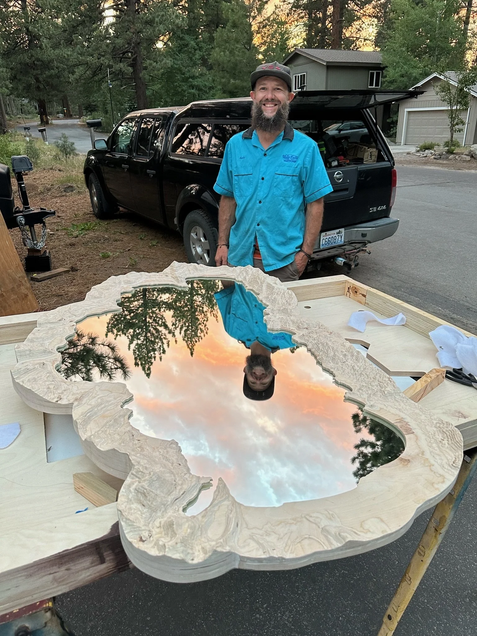 A smiling man with a beard and a black cap standing outdoors beside a table with a wood and resin outdoor art piece reflecting a sunset sky and trees. A black pickup truck and residential houses are in the background.