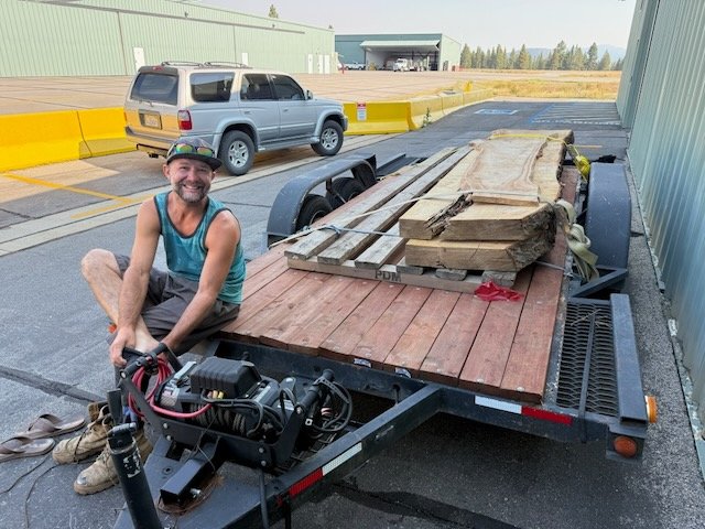 A smiling man in a tank top and shorts sitting on the edge of a flatbed trailer loaded with wooden planks and lumber, parked outside near industrial buildings and a silver SUV, with a clear sky in the background.