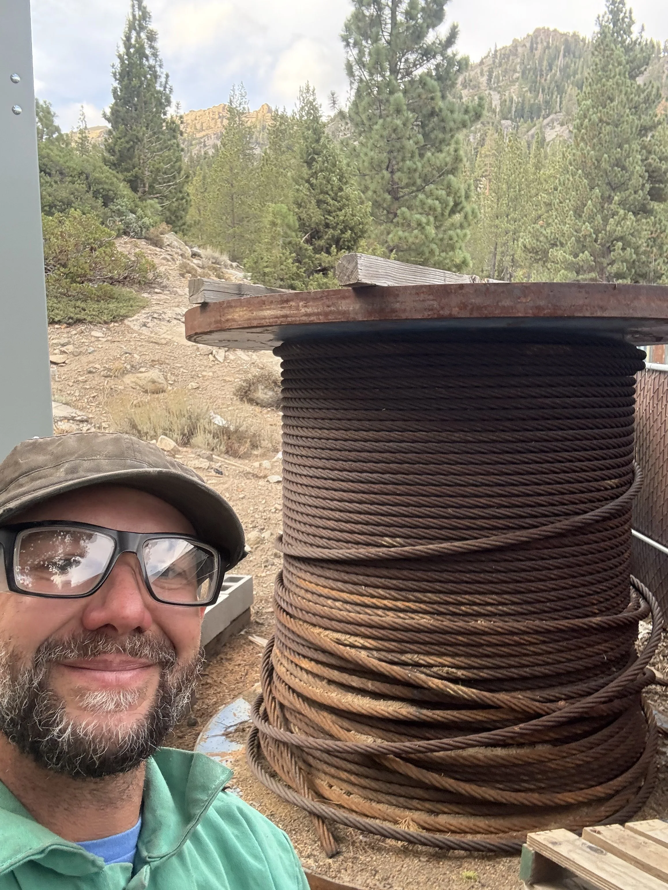 A bearded man wearing glasses, a gray cap, and a green jacket is taking a selfie outdoors in a mountainous area with a large spool of thick, rusty wire in the background. The area is surrounded by trees and rocky terrain.