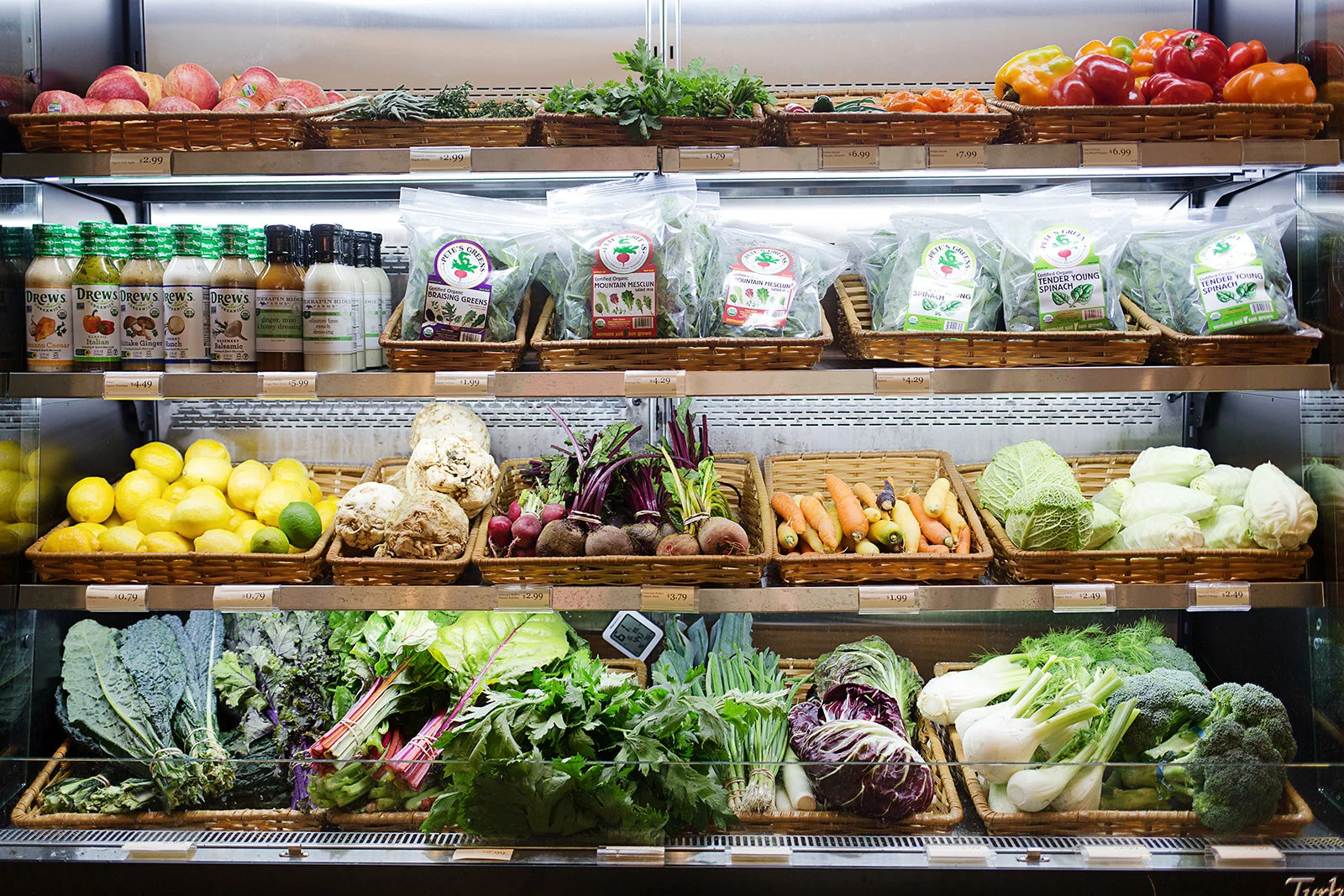 Display of fresh produce in a grocery store, including tomatoes, peppers, apples, lemons, cauliflower, carrots, beets, leafy greens, broccoli, and packaged greens and dressings.