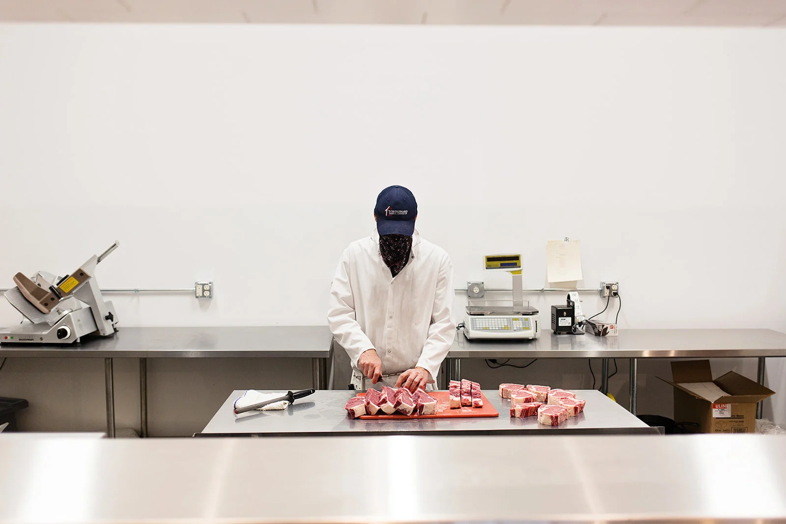 A person in a white coat and face mask, wearing a dark cap, but their face is obscured, is cutting raw meat on a red cutting board in a commercial kitchen with stainless steel counters and equipment.