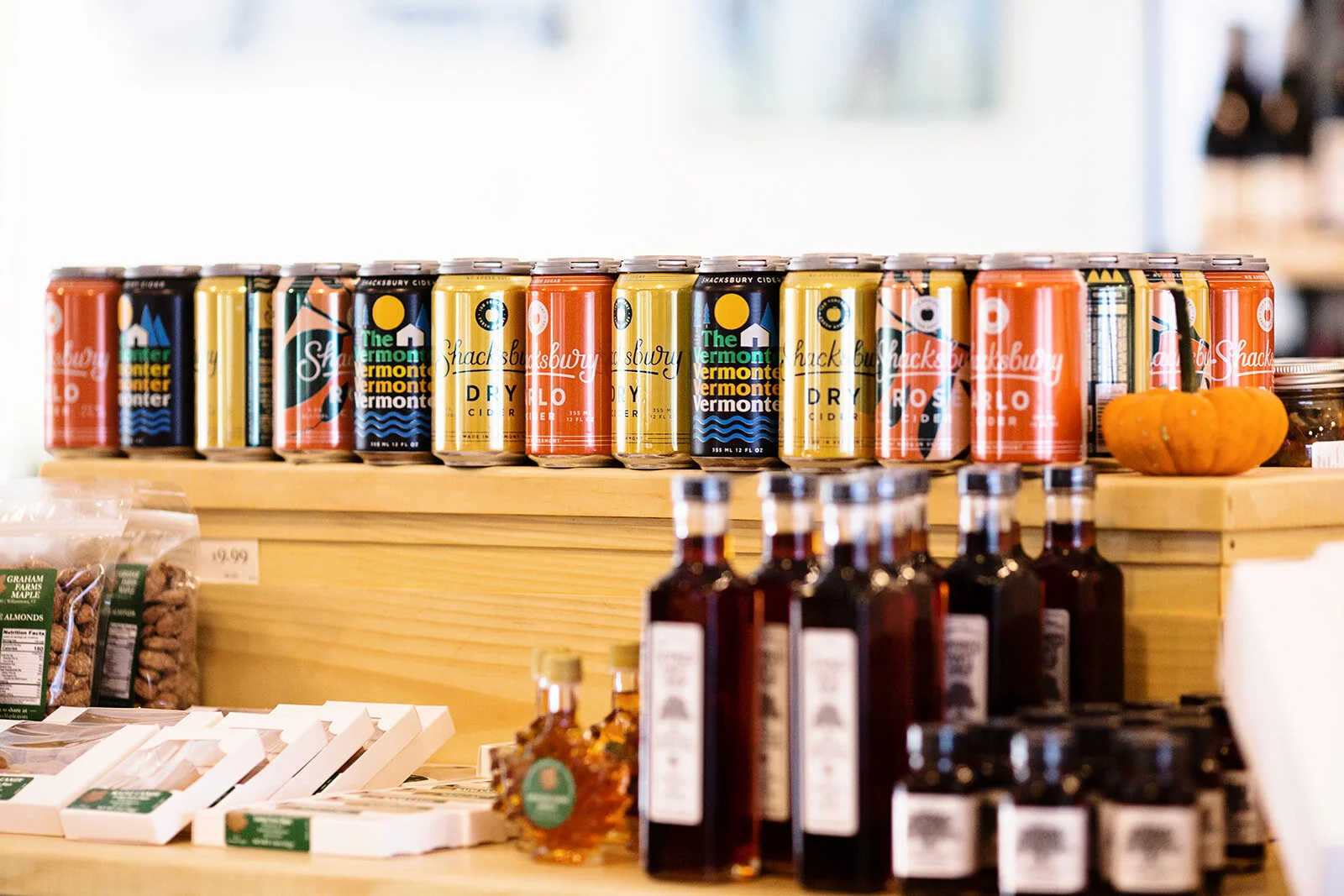 Various colorful cans of Sparkling Vermont Vermonter Dry Cider arranged on a wooden shelf in a store, with small bottles and packaged goods in the foreground.