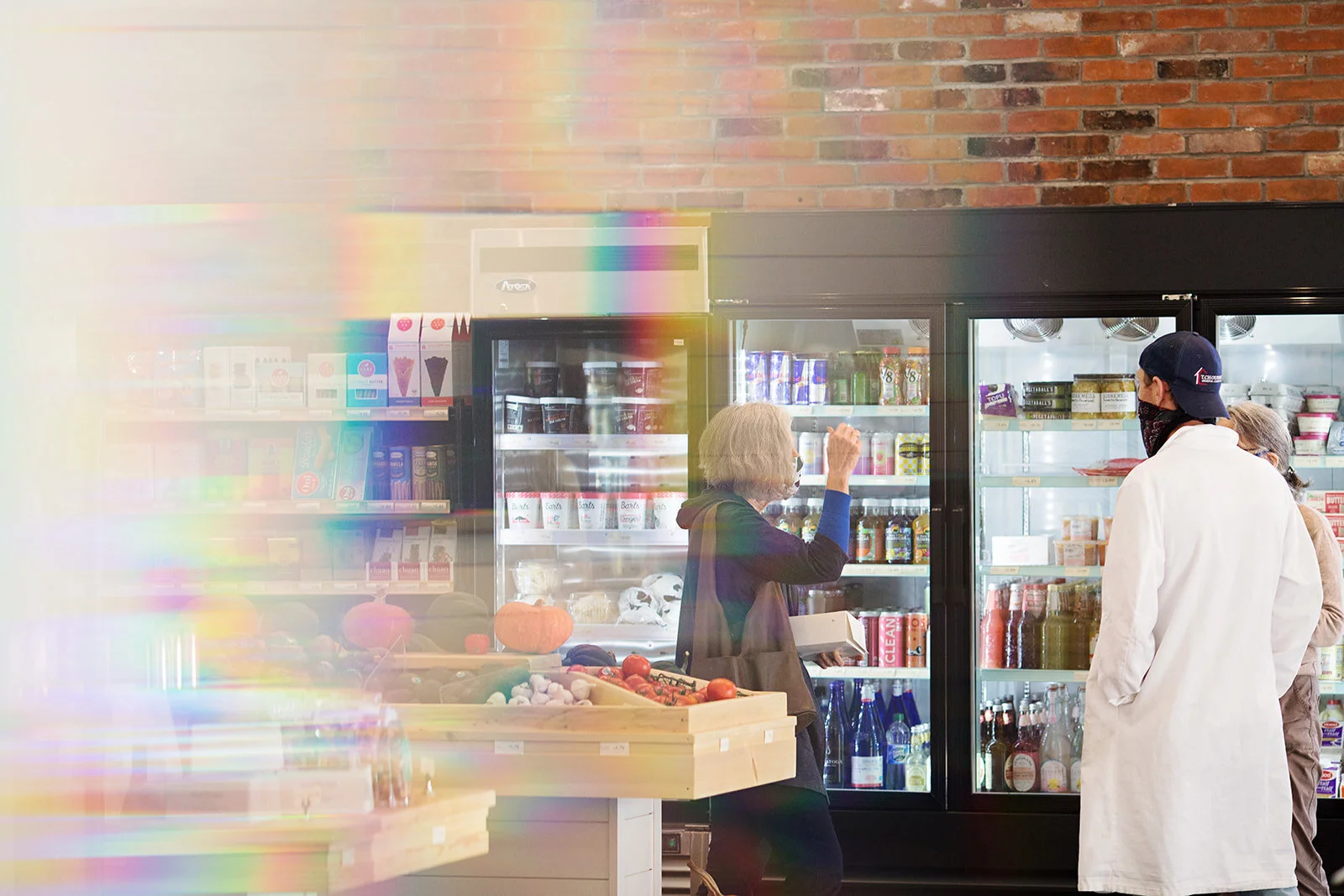 Inside a grocery store, two people are talking near refrigerators filled with drinks and food, with produce on a nearby table and a brick wall in the background.