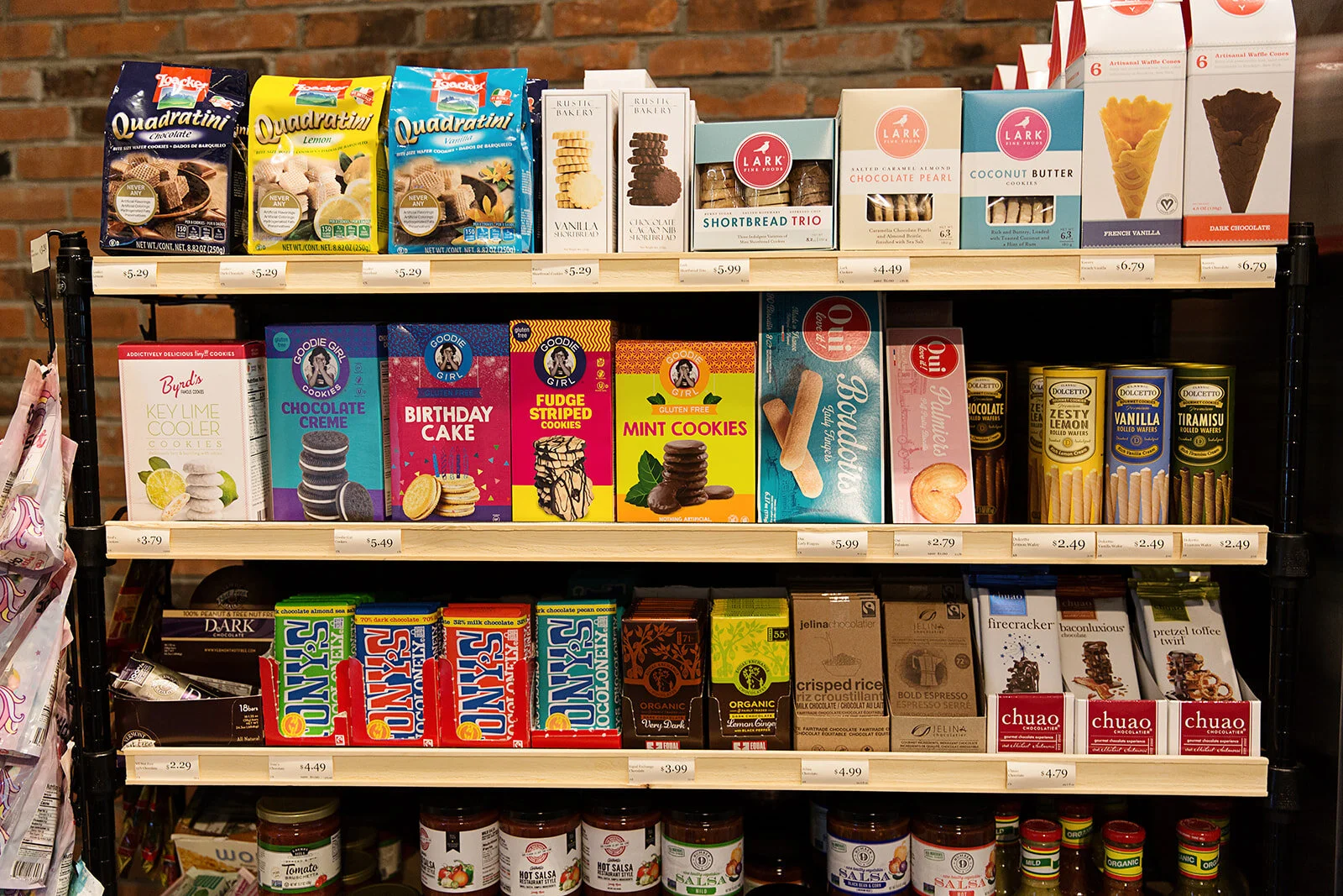 Shelf stocked with various cookies, wafers, and snack items in packages. The top shelf has wafer cookies from Loacker and Lark, priced between $4.49 and $6.79. The middle shelf contains cookie boxes labeled Byrd's, Goodie Girl, and Dolcetto, priced f