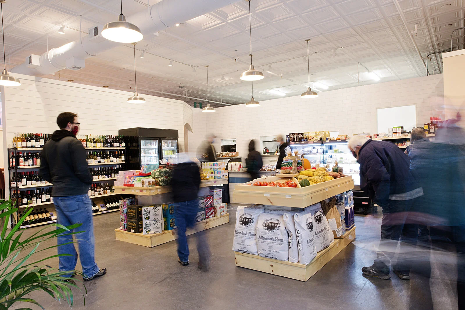 Interior of a grocery store with people shopping, shelves stocked with wine, produce, and other food items, and a refrigerated section in the background.