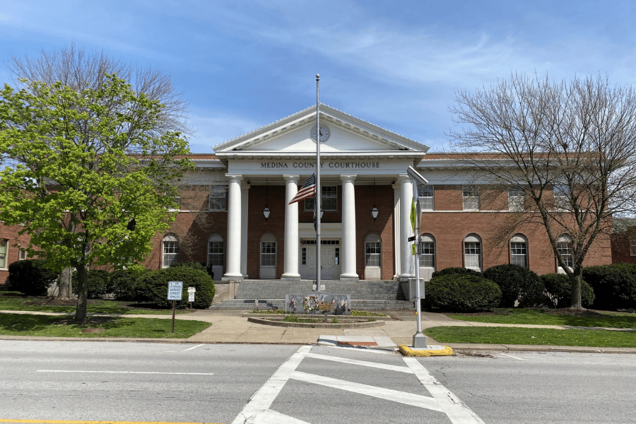 Front view of the Medina County Courthouse in Medina, Ohio, showing the red-brick exterior, white columns, courthouse entrance, and American flag along Public Square.
