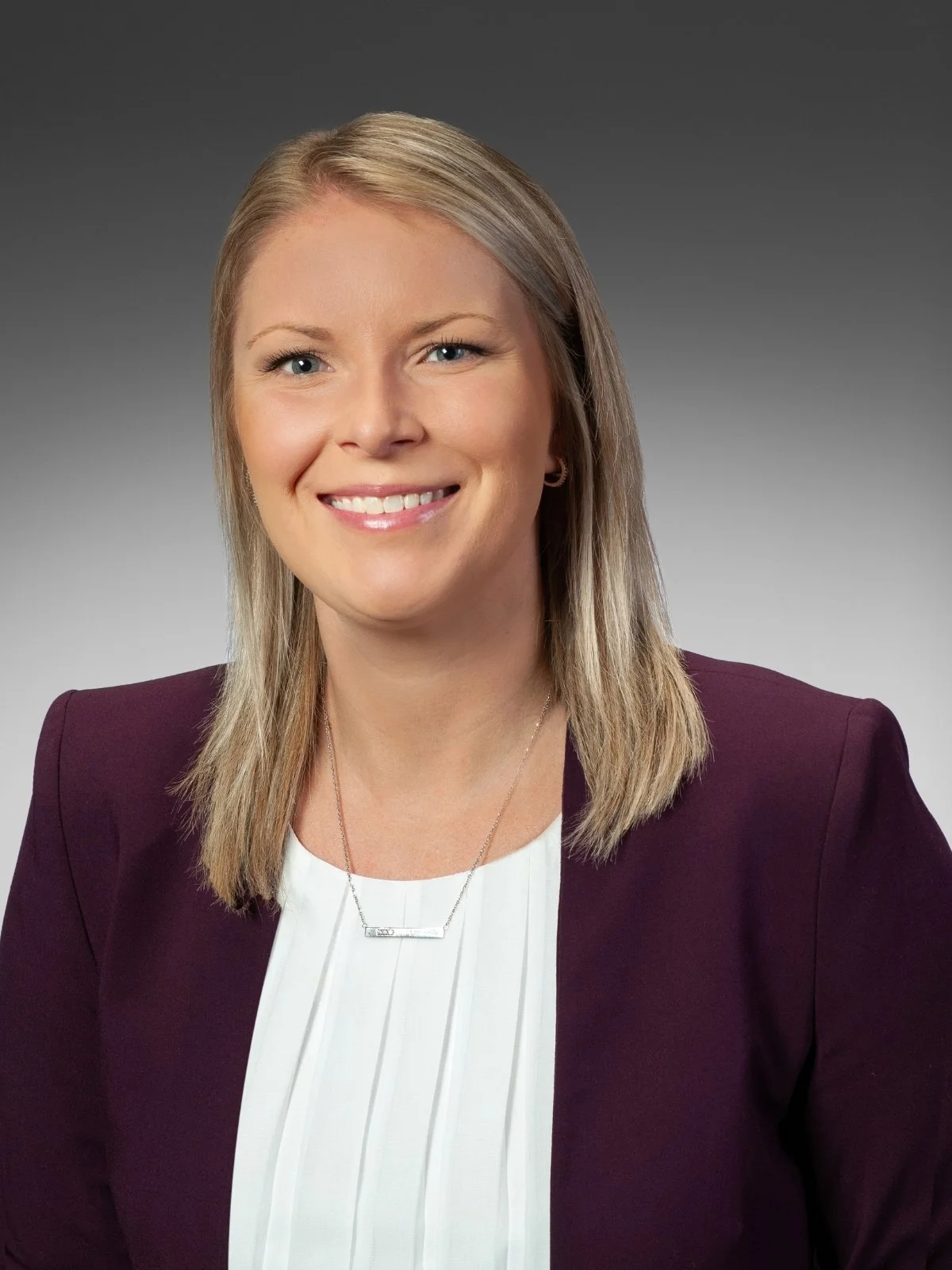 Professional headshot of attorney Alex Lavelle wearing a plum blazer and white blouse against a gray studio background.