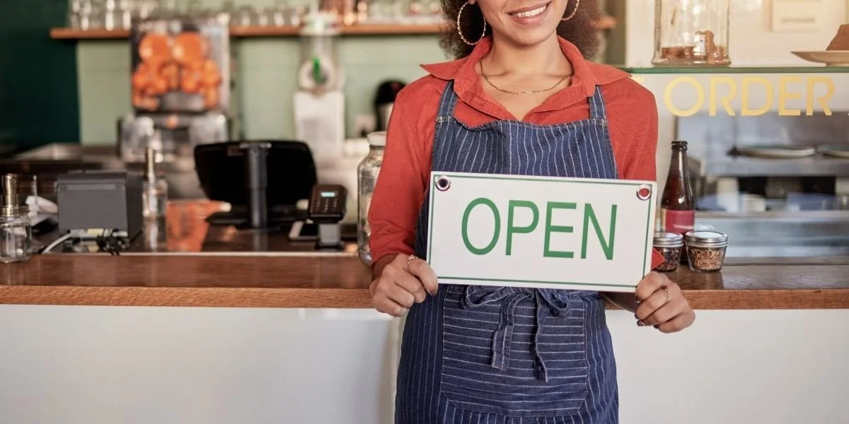 Small business owner standing behind a service counter holding an “Open” sign, representing business operations, growth, and entrepreneurial planning.