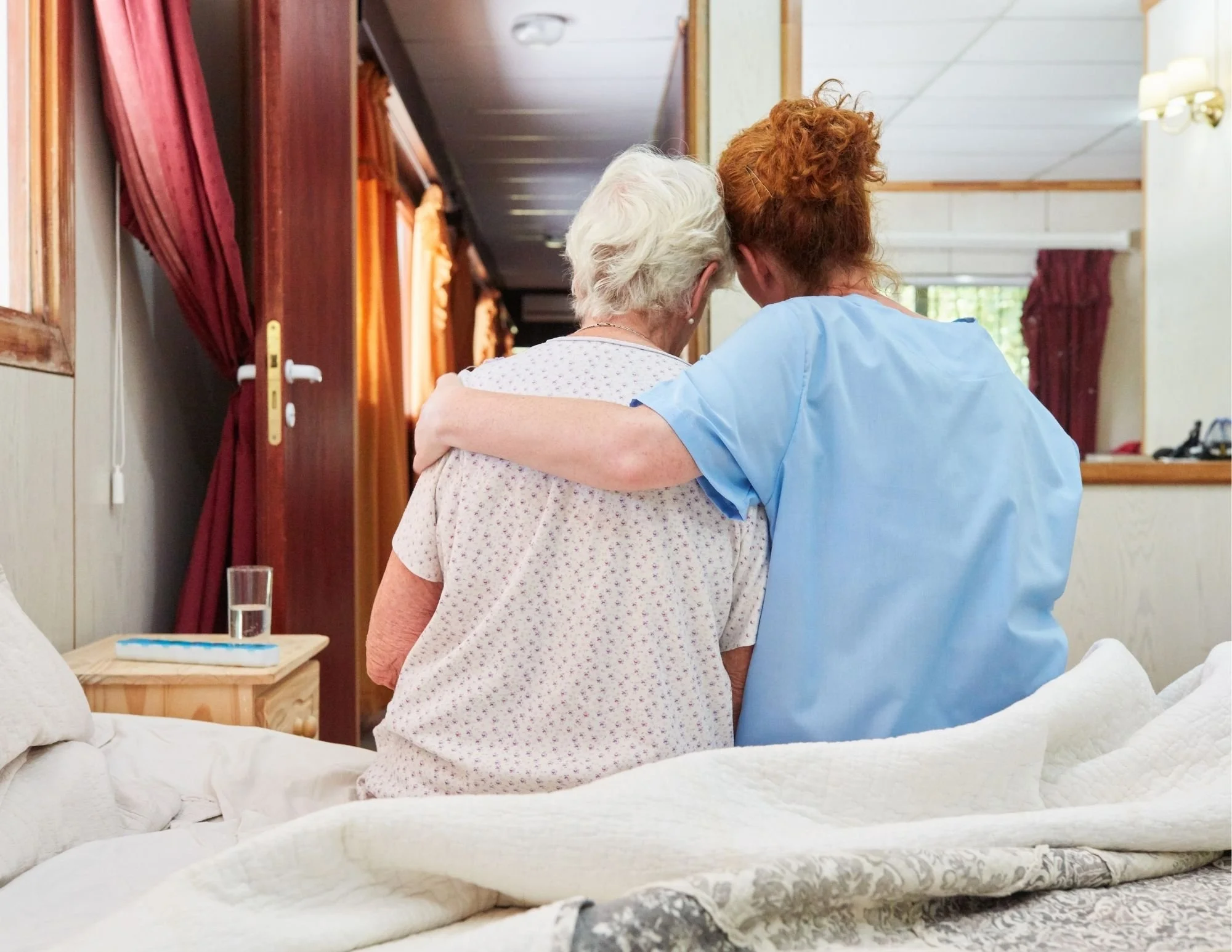 Healthcare professional sitting beside an older adult on a bed, gently placing an arm around their shoulders in a supportive gesture inside a well-lit room with wooden doors and curtains.