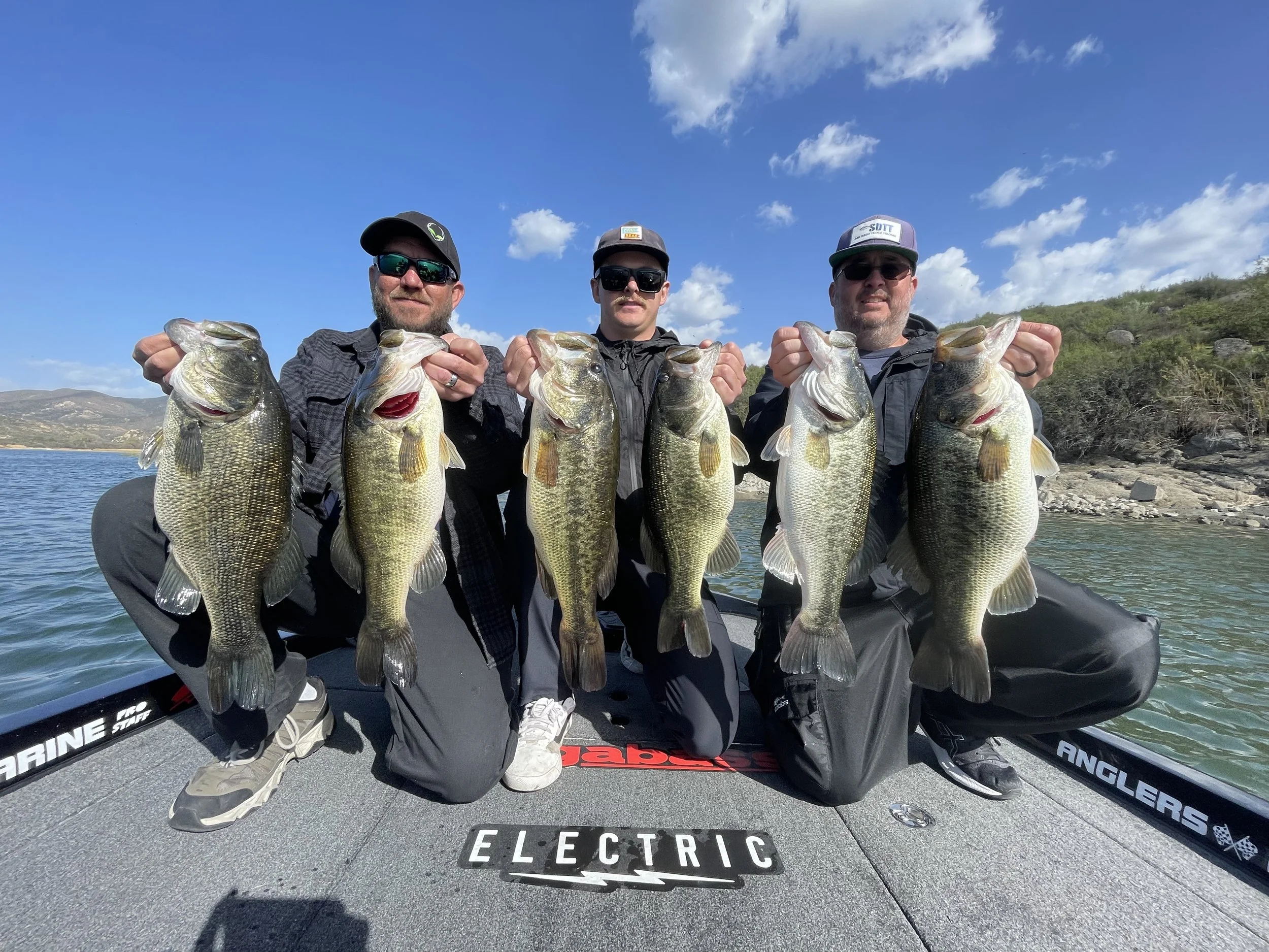Three men kneeling on a boat holding up large fish, with blue sky and hills in the background. Lake skinner Bass Fishing guide running charters and fishing trips.