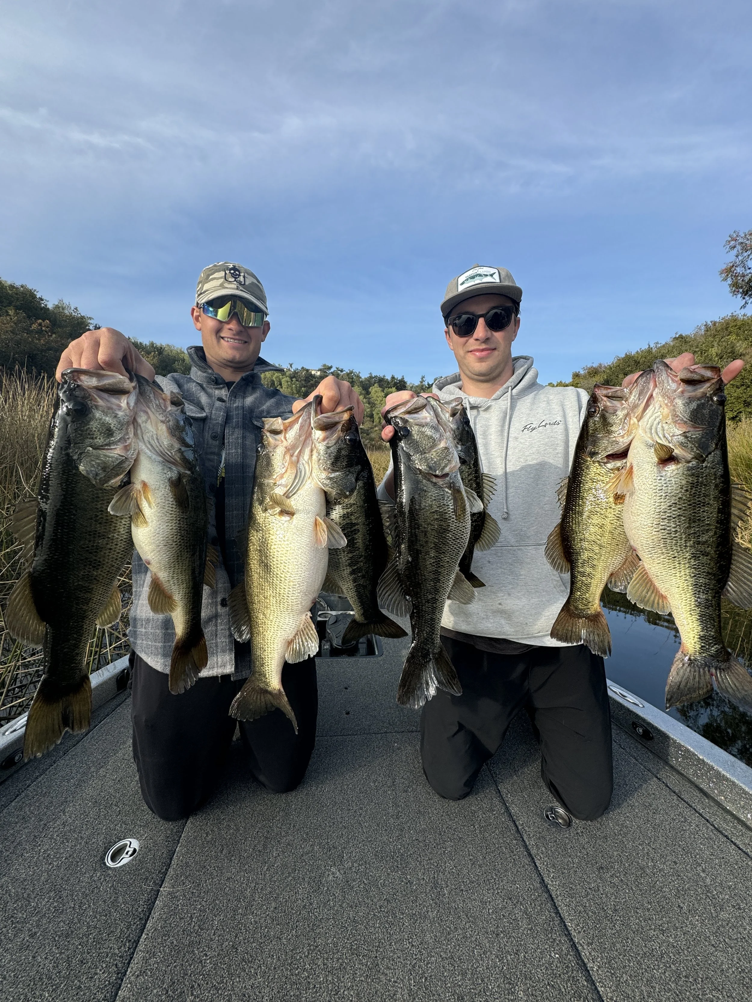 Lake Miramar bass fishing guide San Diego. Two men kneeling on a boat holding five large fish, with water and trees in the background.