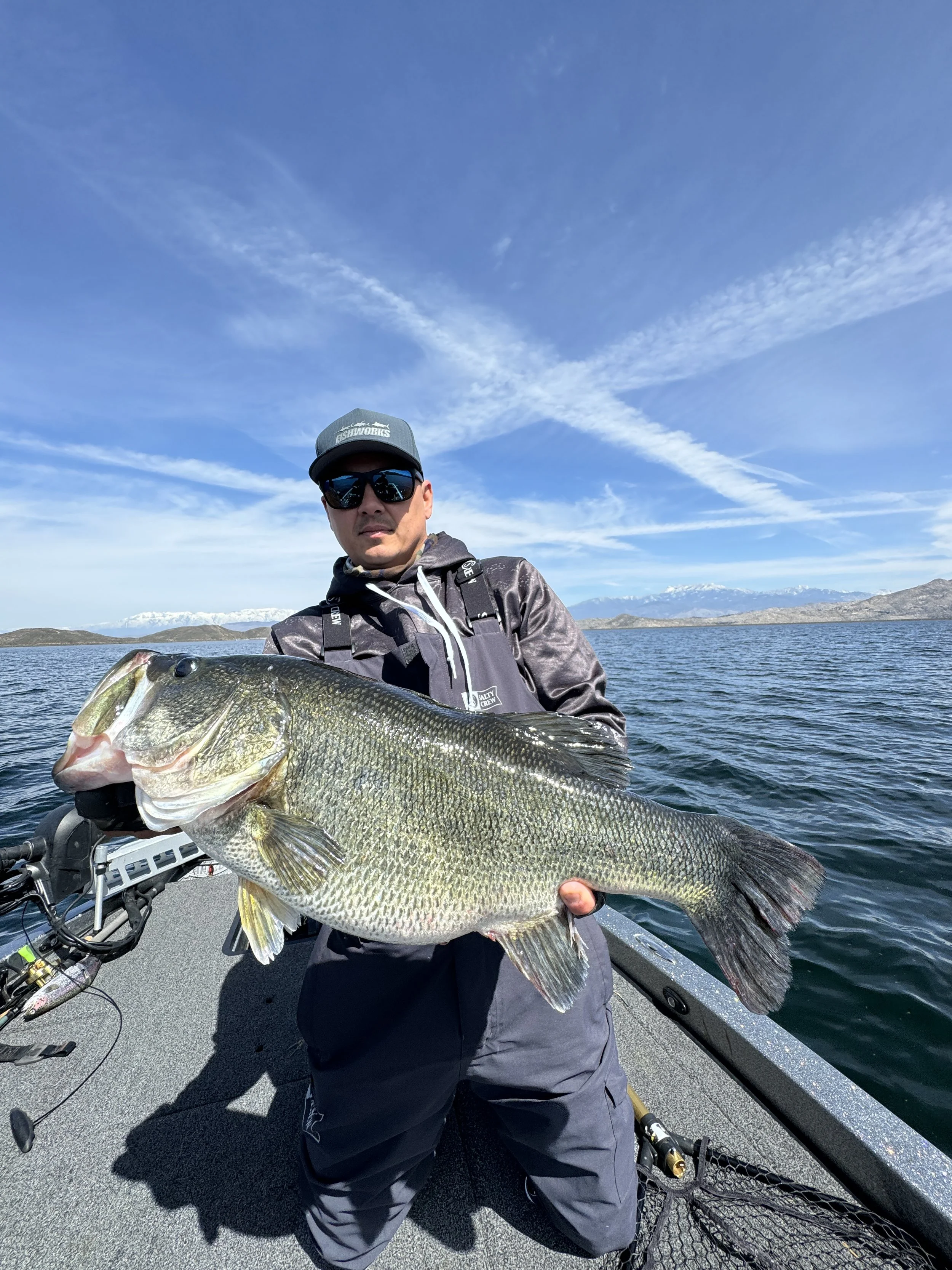 Man in sunglasses and a cap holding a large fish on a boat with water and mountains in the background.