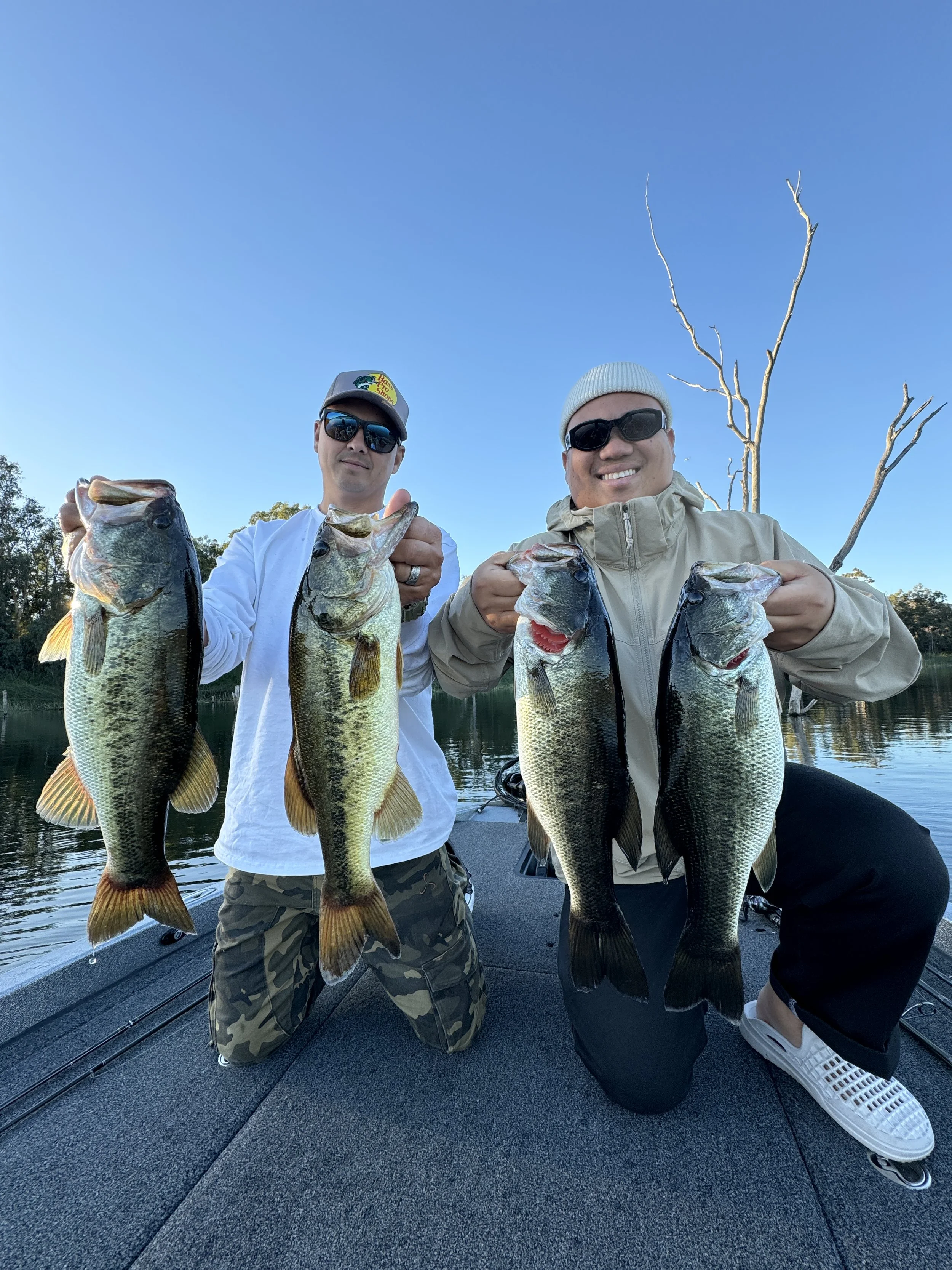 Otay lake  bass fishing guide. Two men kneeling on a boat holding largemouth bass they caught, with a lake and leafless tree in the background on a clear day.