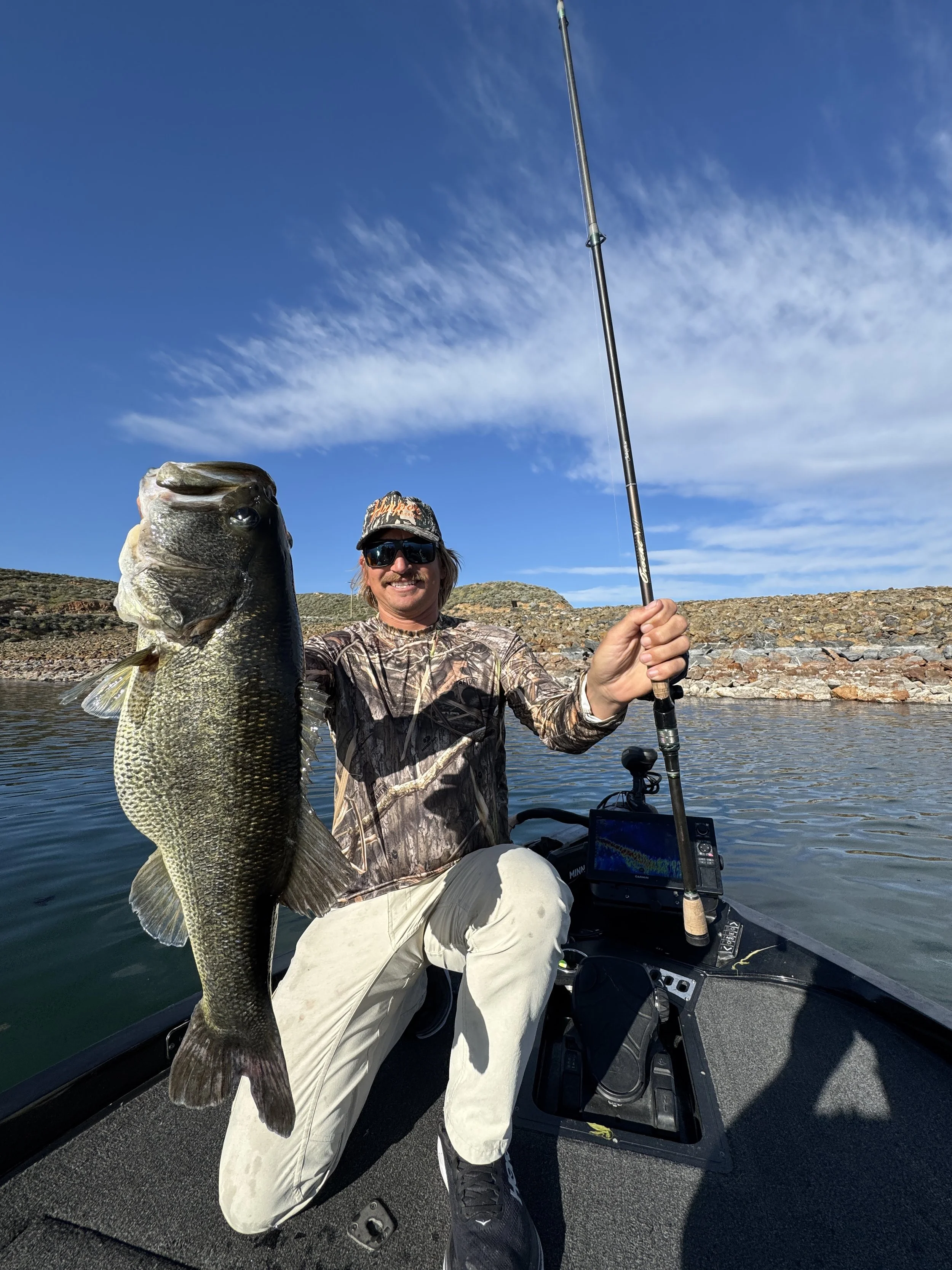 Man in camouflage shirt and sunglasses holding a large fish while sitting in a boat on a lake, with blue sky and rocky shoreline in the background. Diamond valley  Lake Bass Fishing guide running charters and fishing trips.