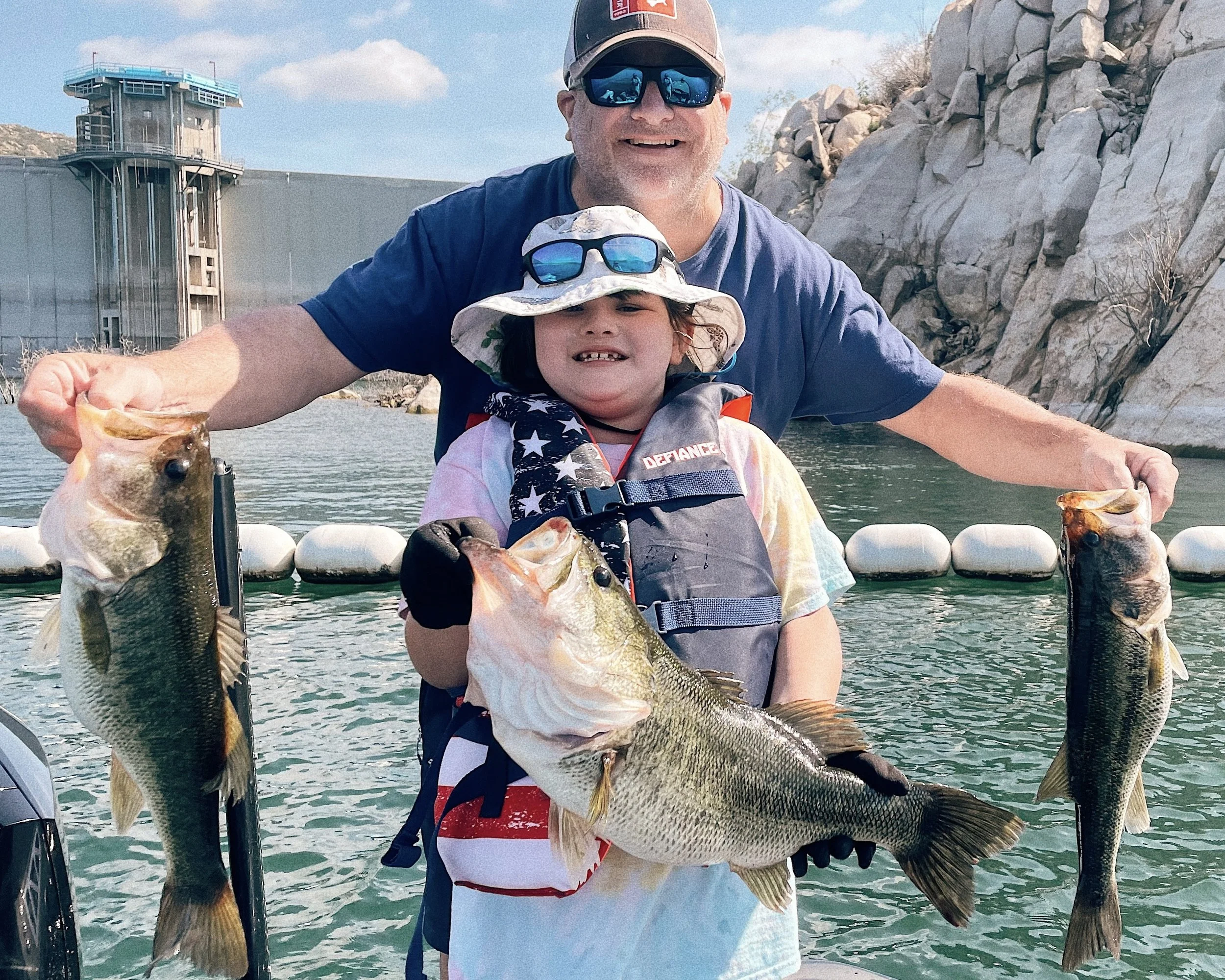A man and a young girl are on a boat in a lake, holding three large fish they caught. The man is smiling and wearing sunglasses and a cap, while the girl wears a sun hat, sunglasses, and a life jacket. The background shows rocky terrain and a dam.