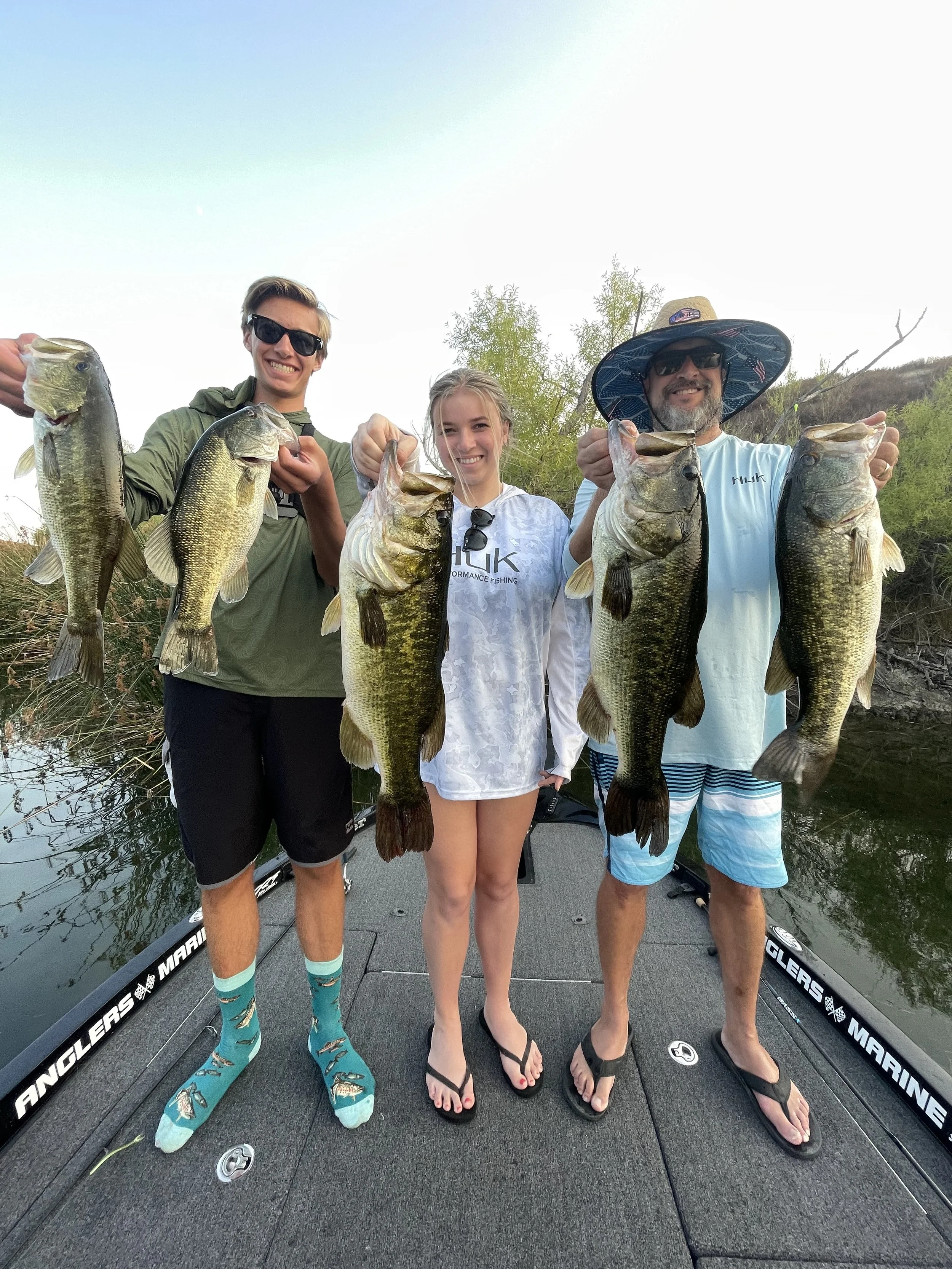Three people on a boat holding large bass fish they caught, with water and trees in the background. Lake skinner Bass Fishing guide running charters and fishing trips.