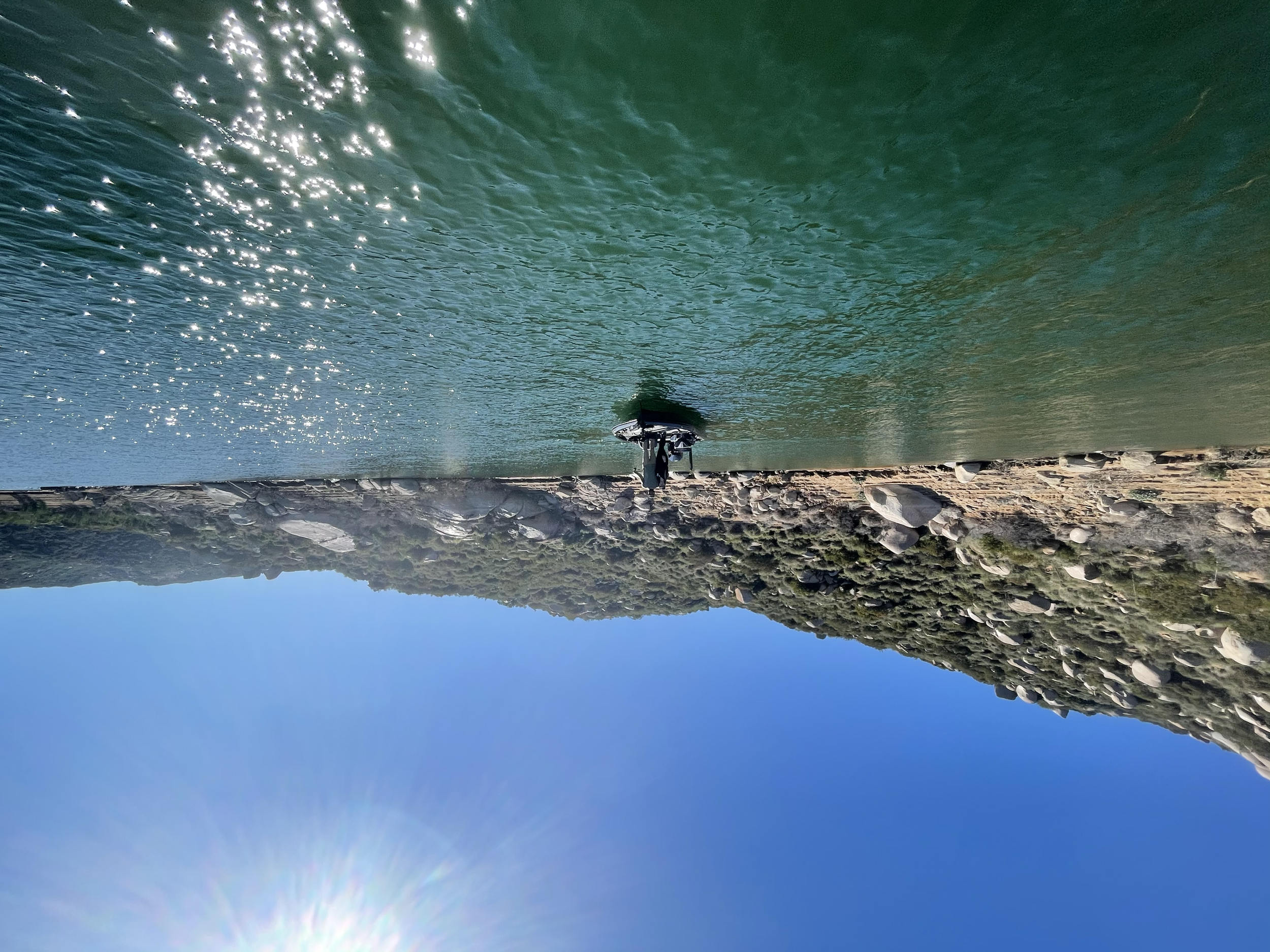 A boat floating on a calm river with a rocky shoreline and hilly terrain in the background under a clear blue sky and bright sunlight.