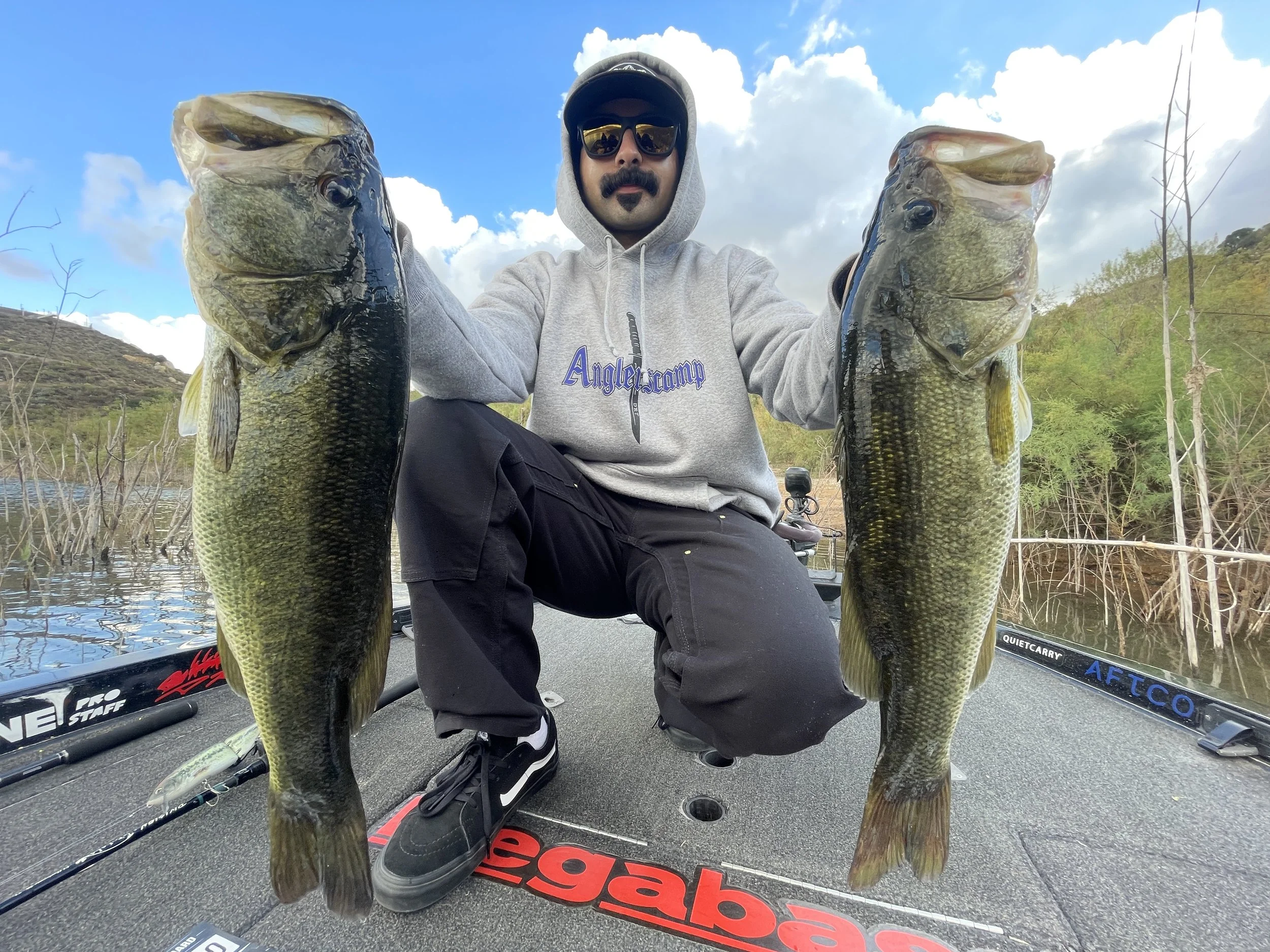 Man wearing sunglasses and a hoodie kneeling on a boat, holding two large bass fish, one in each hand, with a lake and wooded hills in the background.San vincente reservoir bass fishing guide in souther California running trips and charters.