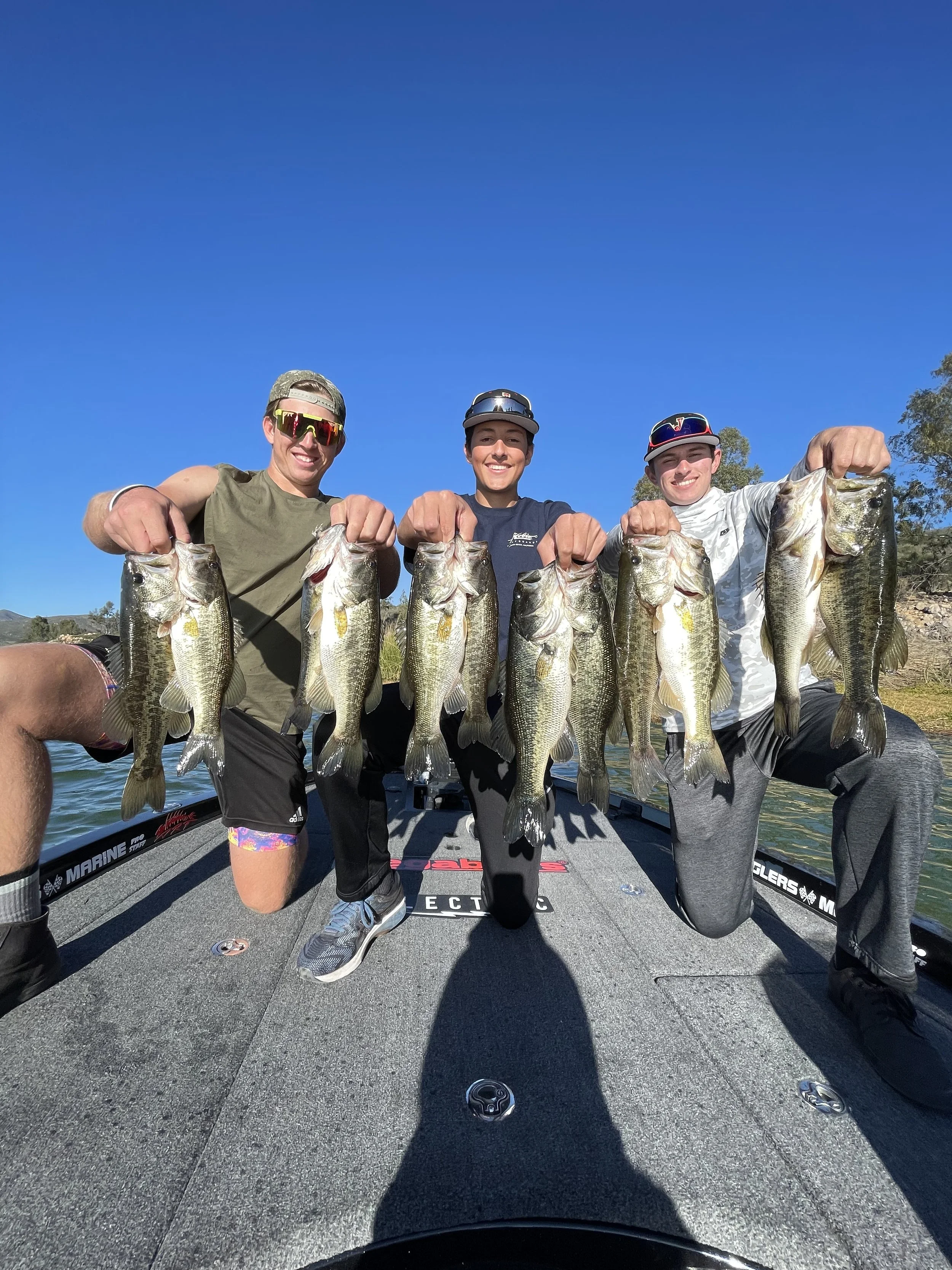 Otay lake bass fishing guide San Diego. Four people holding up largemouth bass fish on a boat, smiling under a clear blue sky.