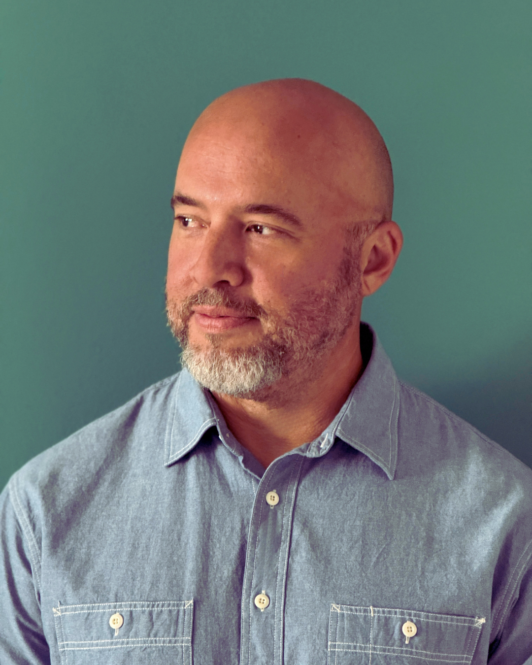 A bald man with a beard wearing a denim button-up shirt, looking to his left, against a plain green background.