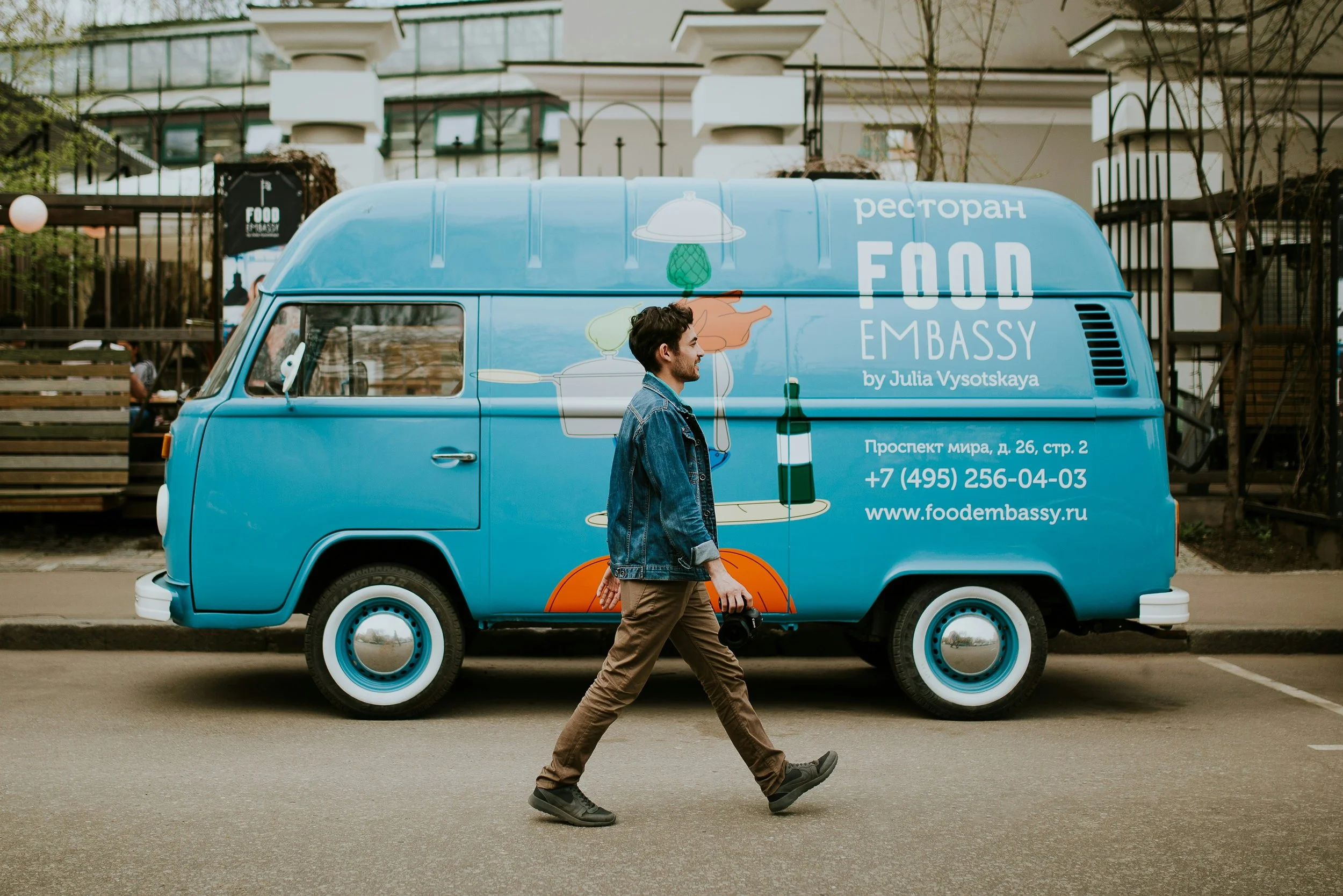 A man walking across a street with a black camera in his right hand, passing by a blue food truck with colorful graphics and Cyrillic writing, in an urban setting with buildings and trees in the background.