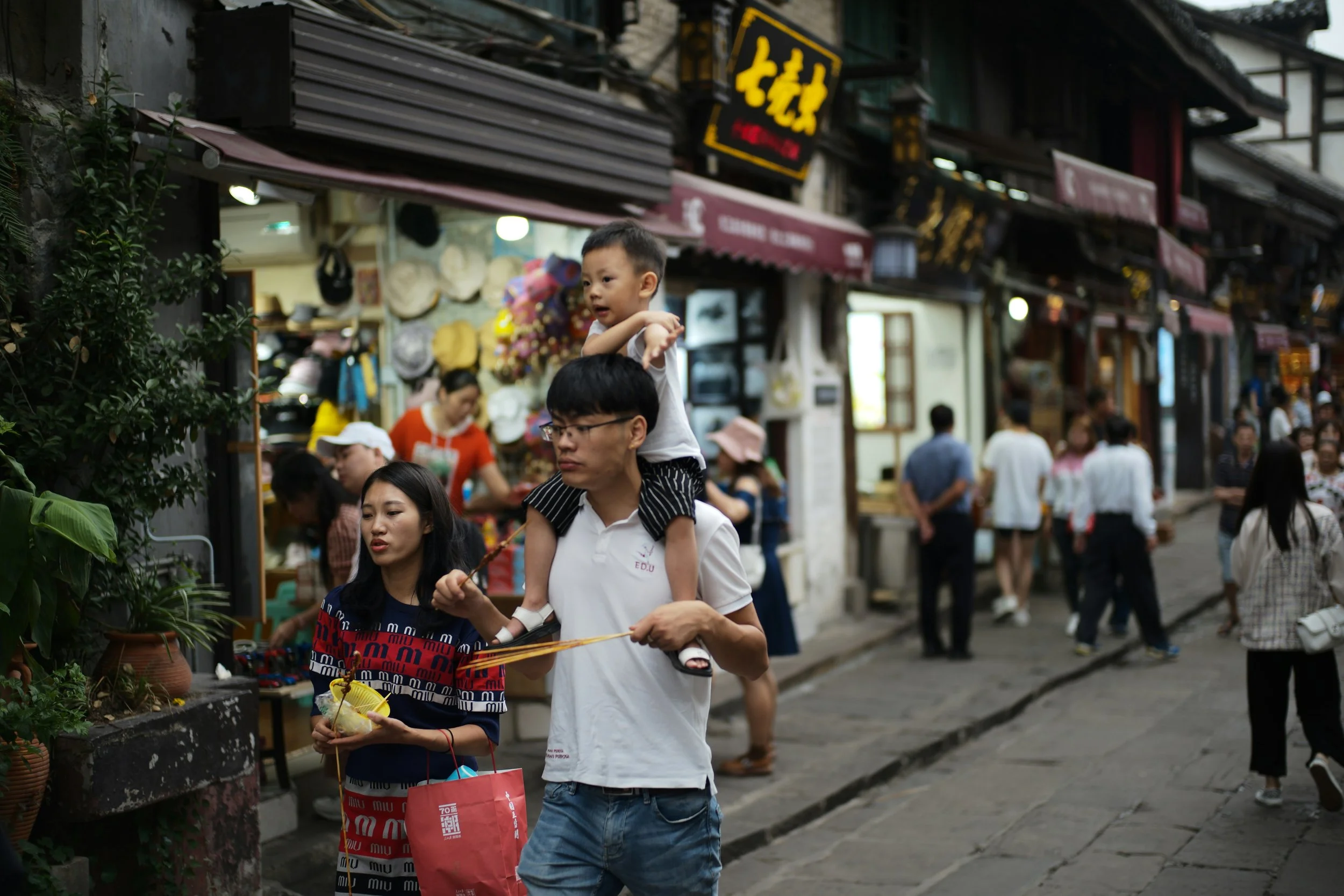 Crowded street market with people shopping, including a man carrying a child on his shoulders, and various store signs in an Asian script.