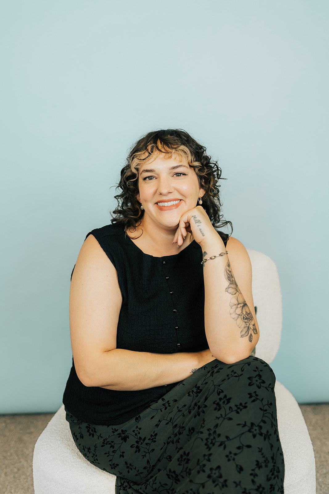 Woman with dark curly hair sitting smiling in a black tank top and checkered skirt