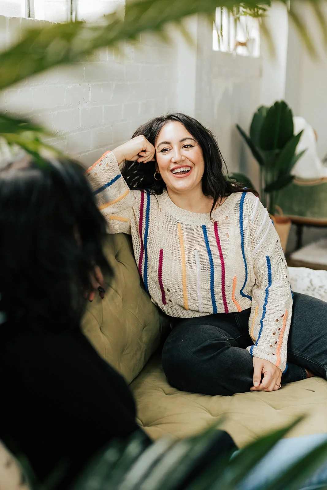 A woman seated on a green couch, wearing a colorful striped sweater, smiling and talking to another person, with plants in the foreground."