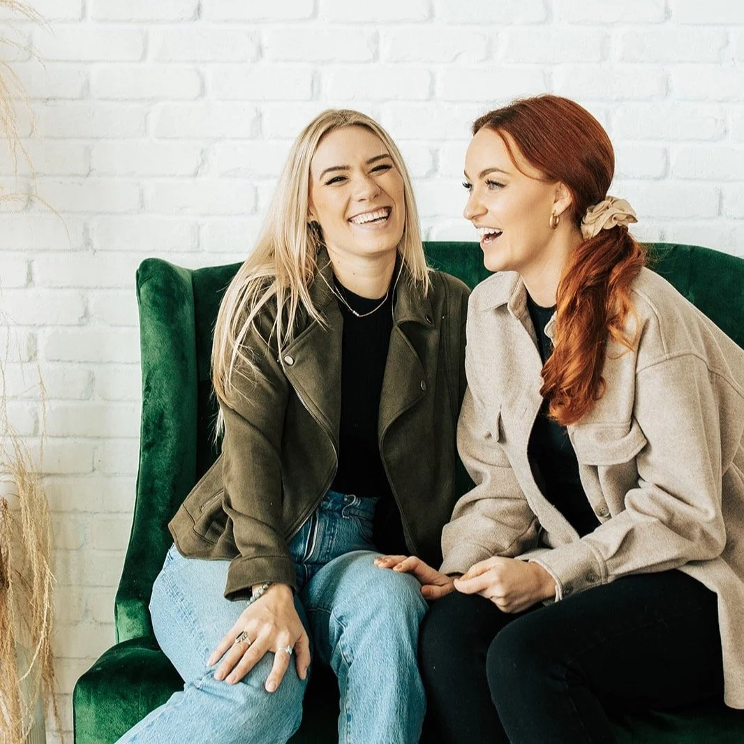 Two women sitting on a green sofa, smiling and laughing in front of a white brick wall. One has blonde hair and is wearing a green jacket and jeans, the other has red hair and is wearing a beige jacket and black pants.