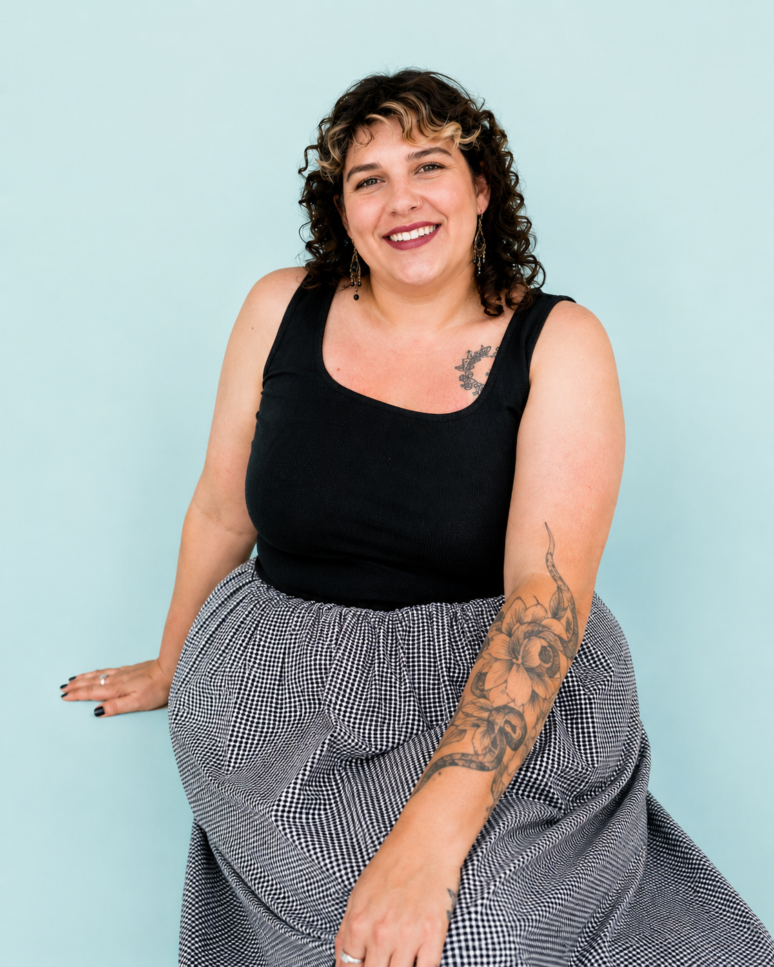 Darked curly haired woman sitting in black tank top and checkered print skirt. 
