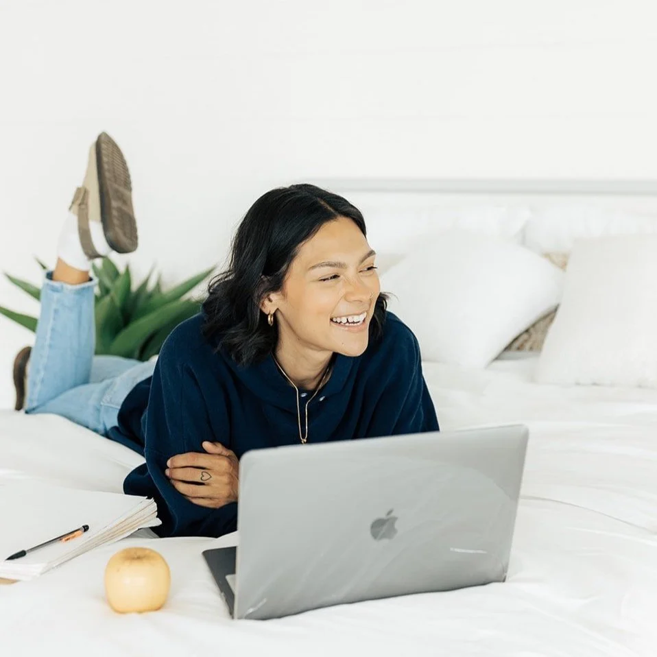 Person lying on a bed using a laptop, smiling. There are notebooks, a pen, and an apple nearby.