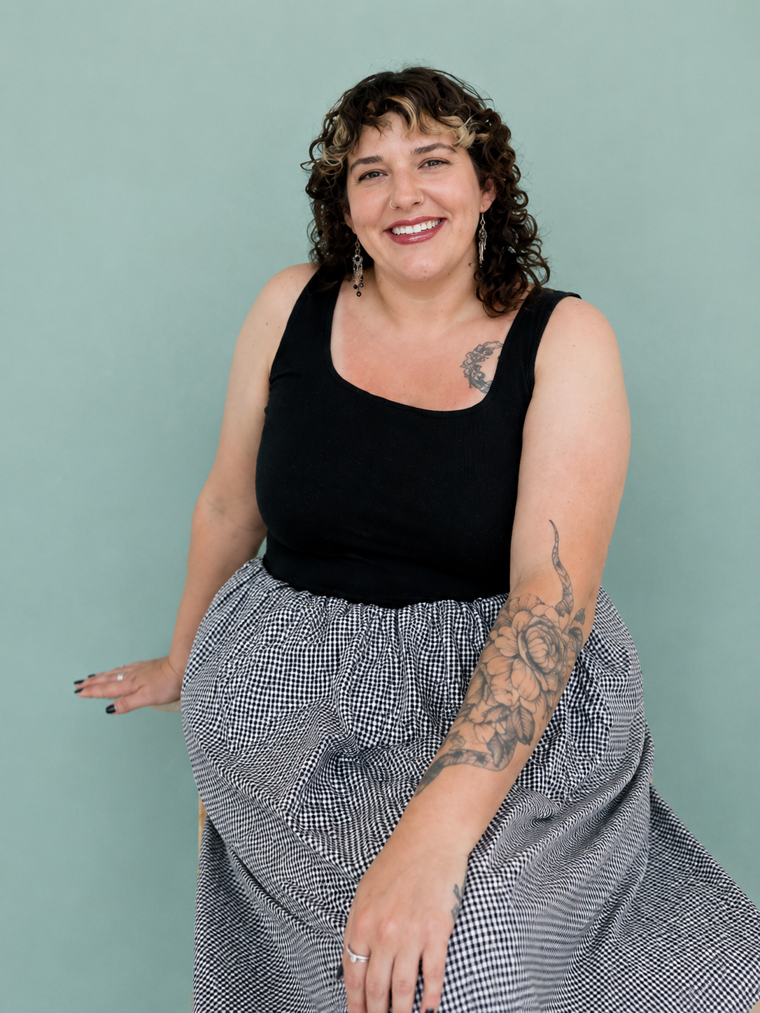Woman with dark curly hair sitting smiling in a black tank top and checkered skirt