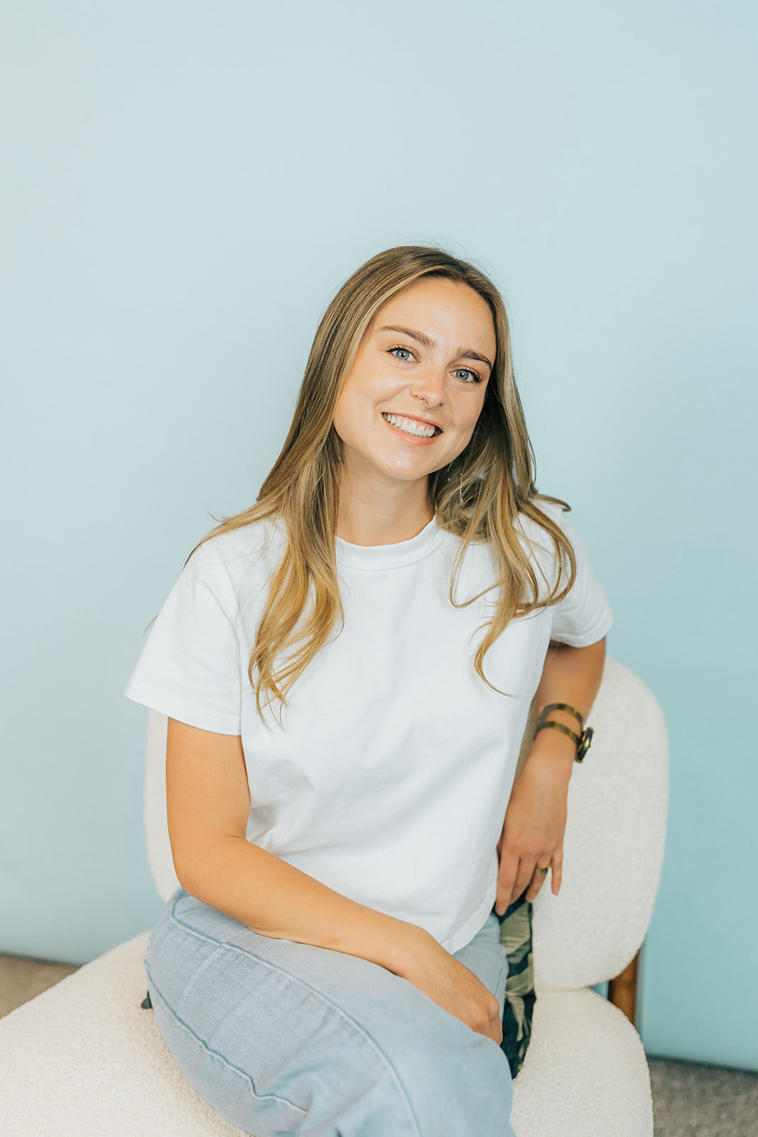 Blonde woman sitting in chair on light blue background wearing a white t shirt and light blue jeans