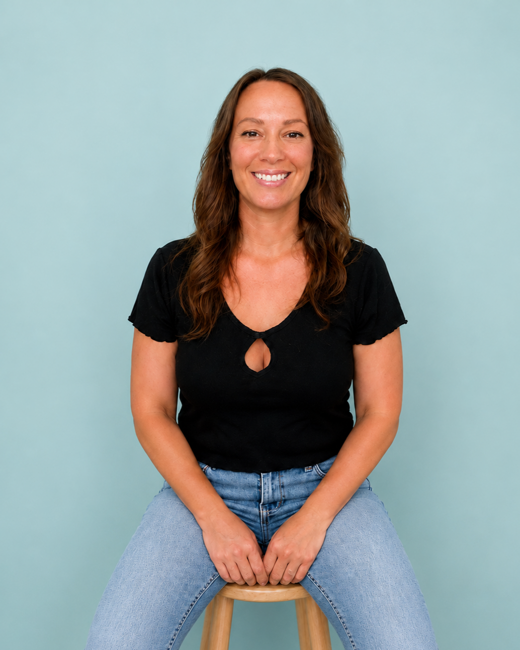 Smiling dark haired woman sitting on stool in black shirt and blue jeans