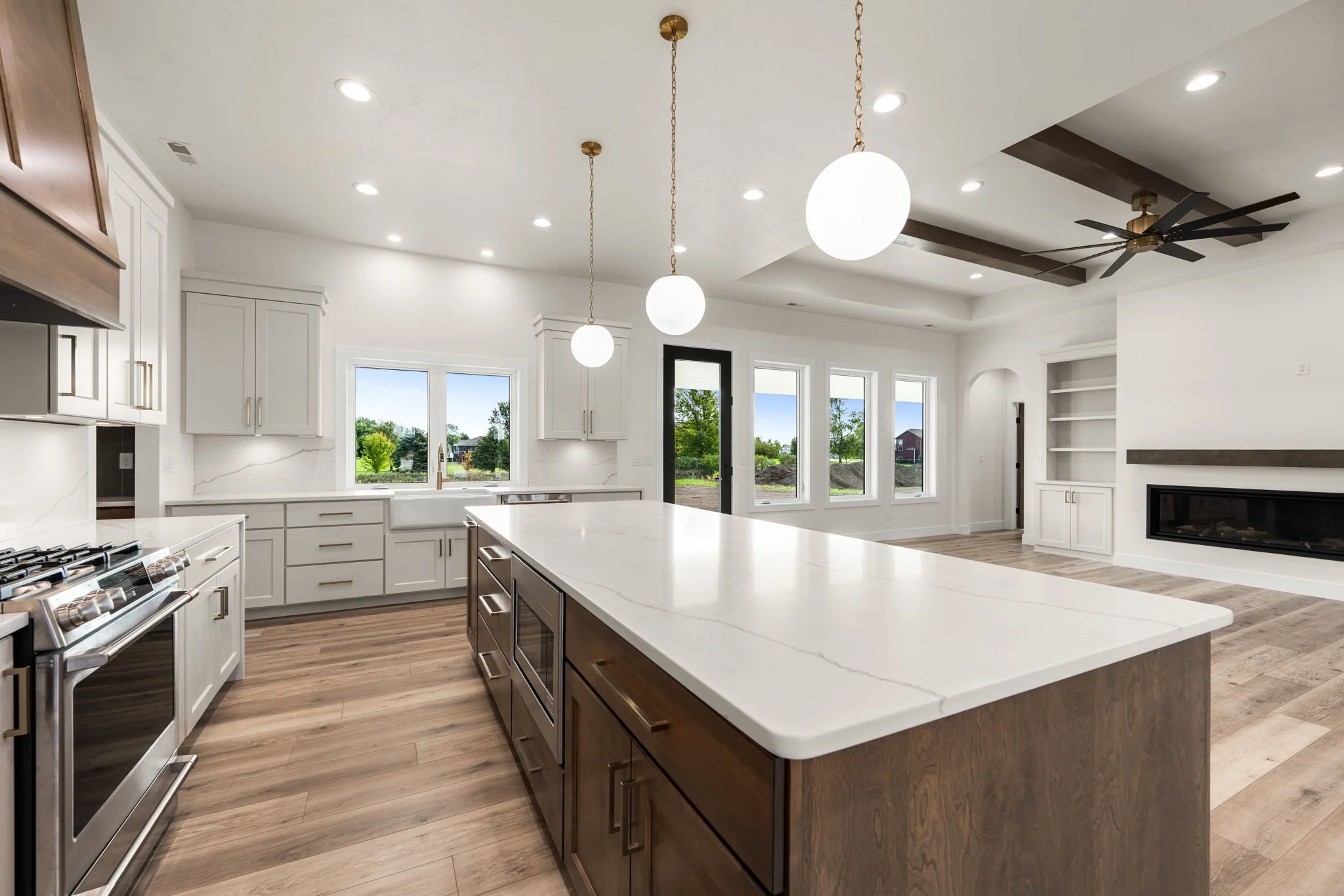 Modern open-concept kitchen with white cabinetry, a large island with dark wood cabinets, pendant lights, stainless steel appliances, a window with a scenic view, and a ceiling fan.
