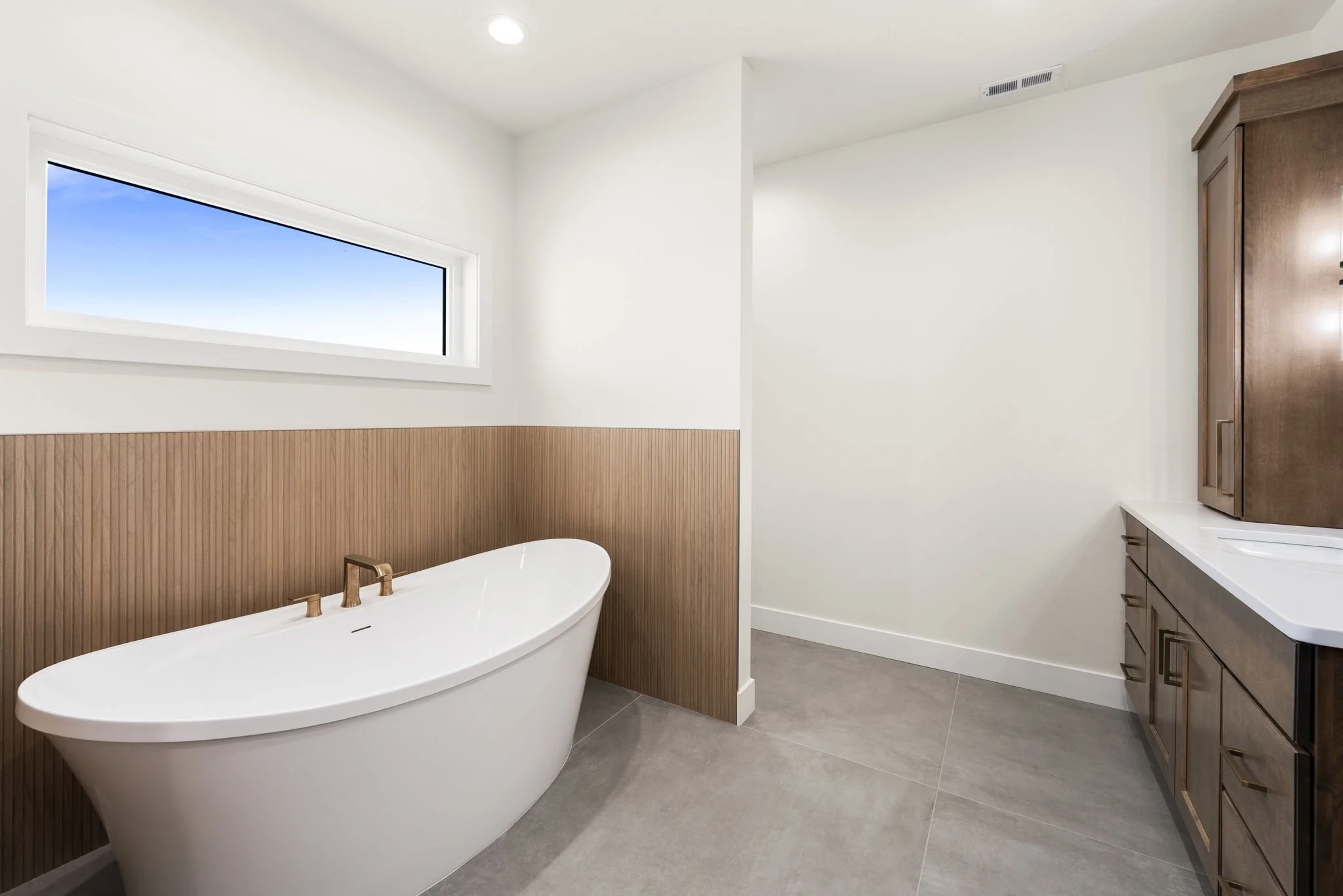 Modern bathroom with white freestanding bathtub, wood paneling on the lower half of the walls, a large horizontal window showing a blue sky, and gray tile flooring. There is a wooden cabinet and a white countertop with a sink.