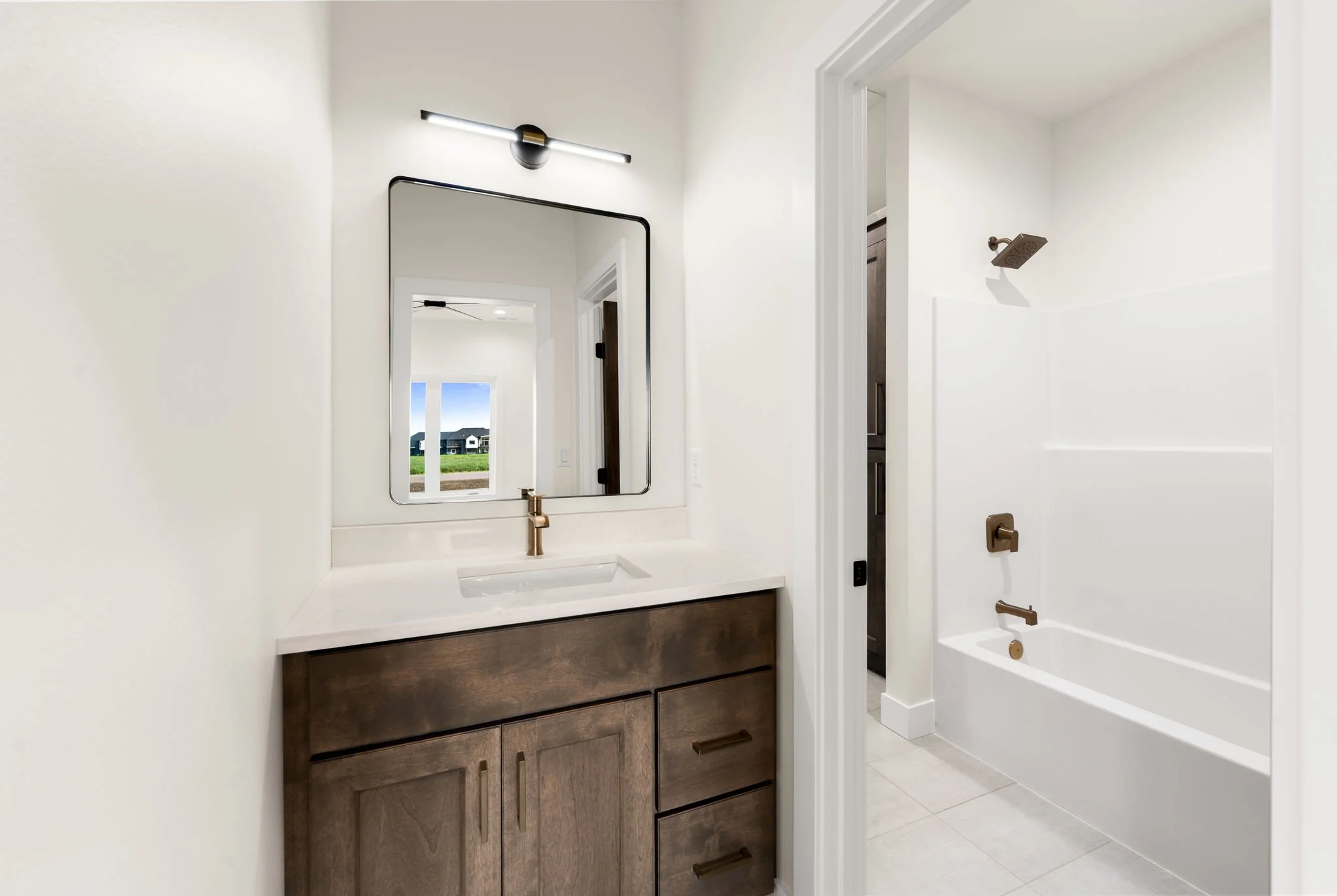 Bathroom with modern dark wood vanity, white countertop, mirror, gold faucet, shower with rain shower head, white walls, door open to a bedroom with a window showing a view of green grass and houses.