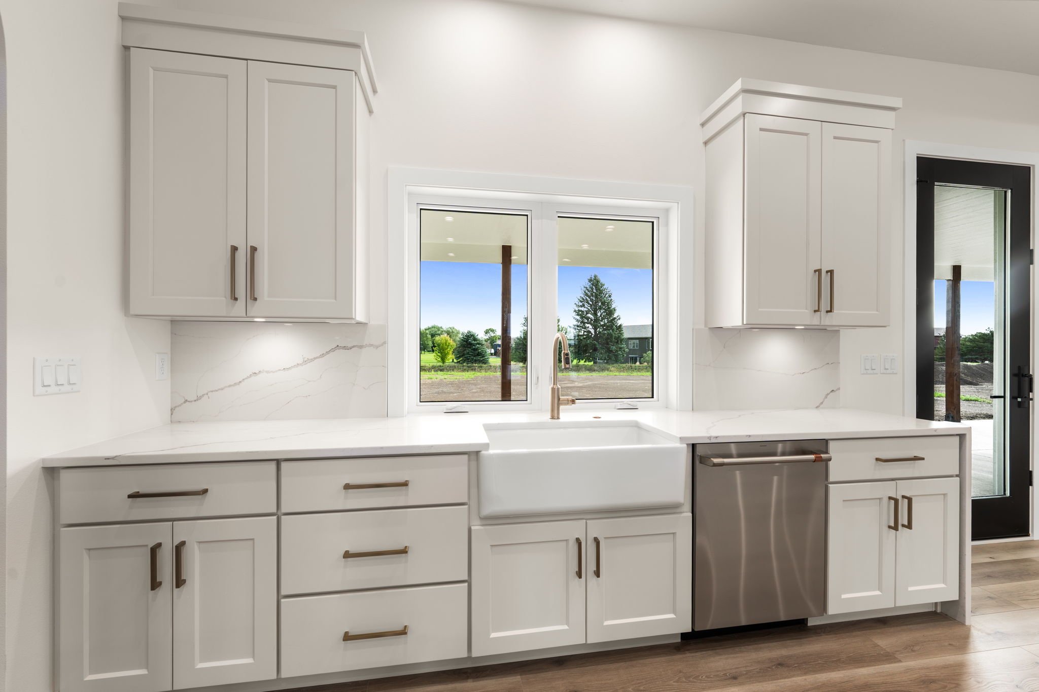 Modern white kitchen with cabinets, marble countertops, window view of green trees, and stainless steel dishwasher.