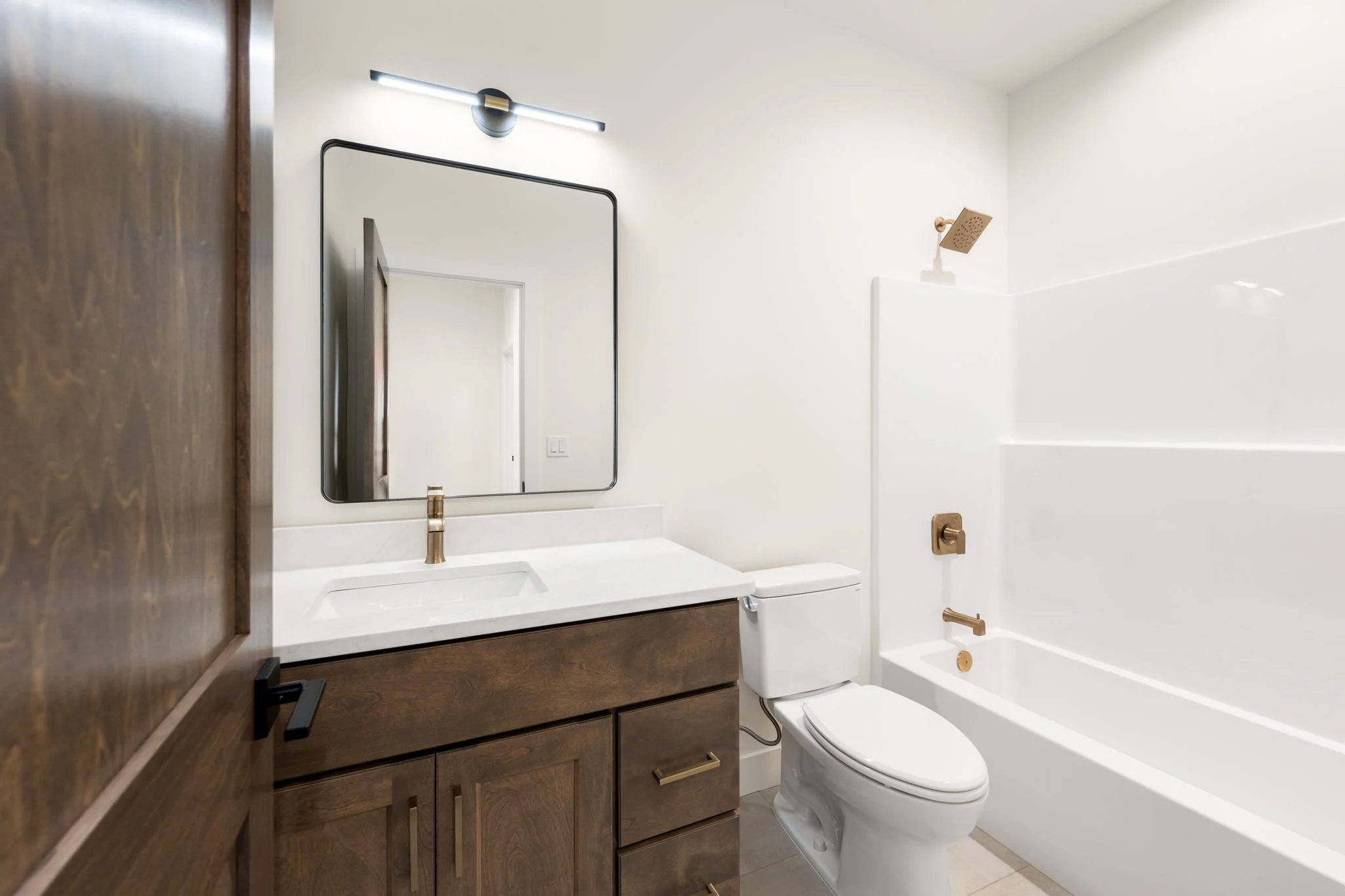 Modern bathroom with white walls, a large mirror, a gold faucet, a wooden vanity, a toilet, a bathtub with a gold faucet, and a showerhead.