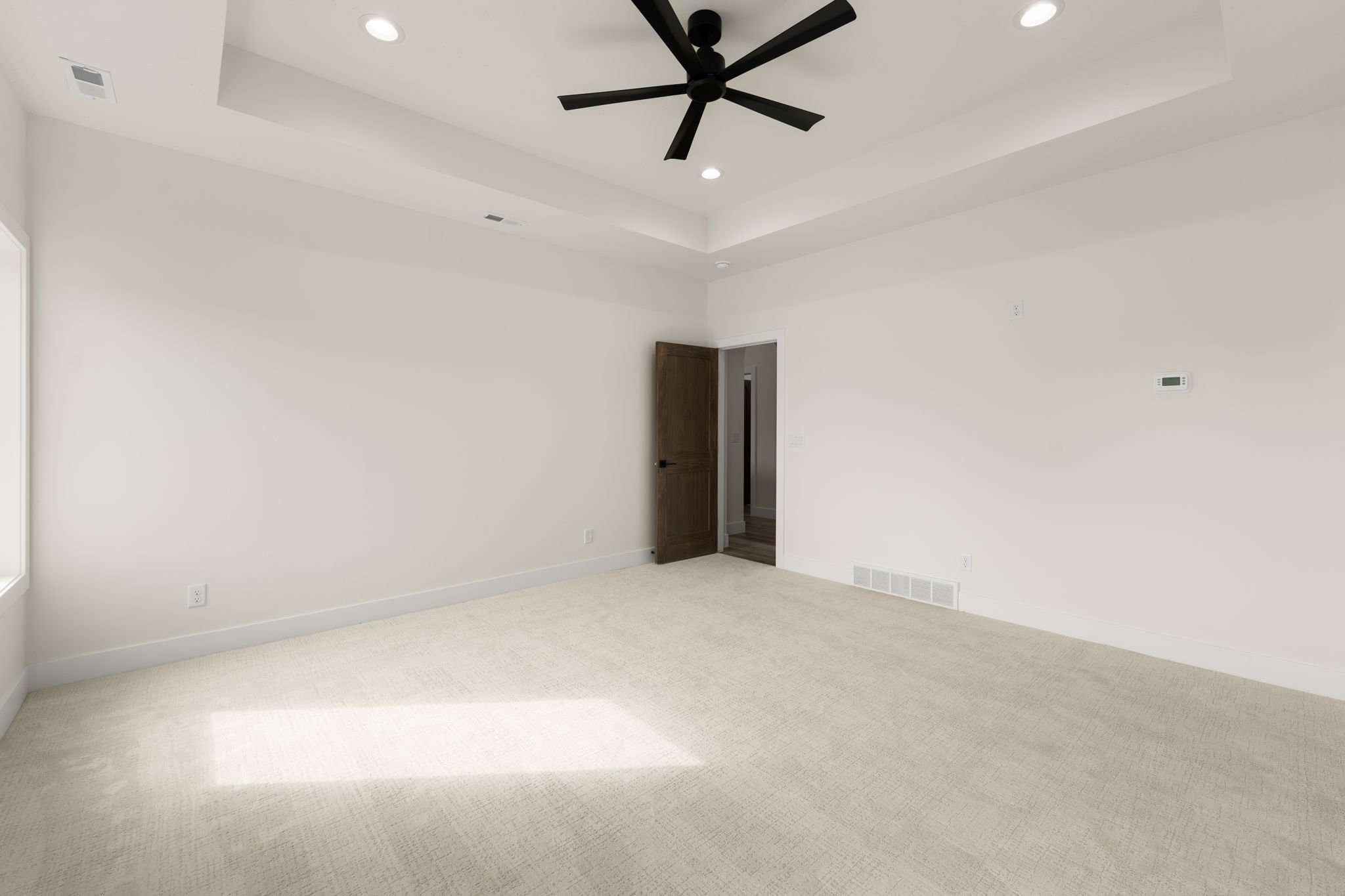 Empty room with cream-colored carpet, white walls, wood door slightly open, ceiling fan, recessed lights, small vents, thermostat on the wall.